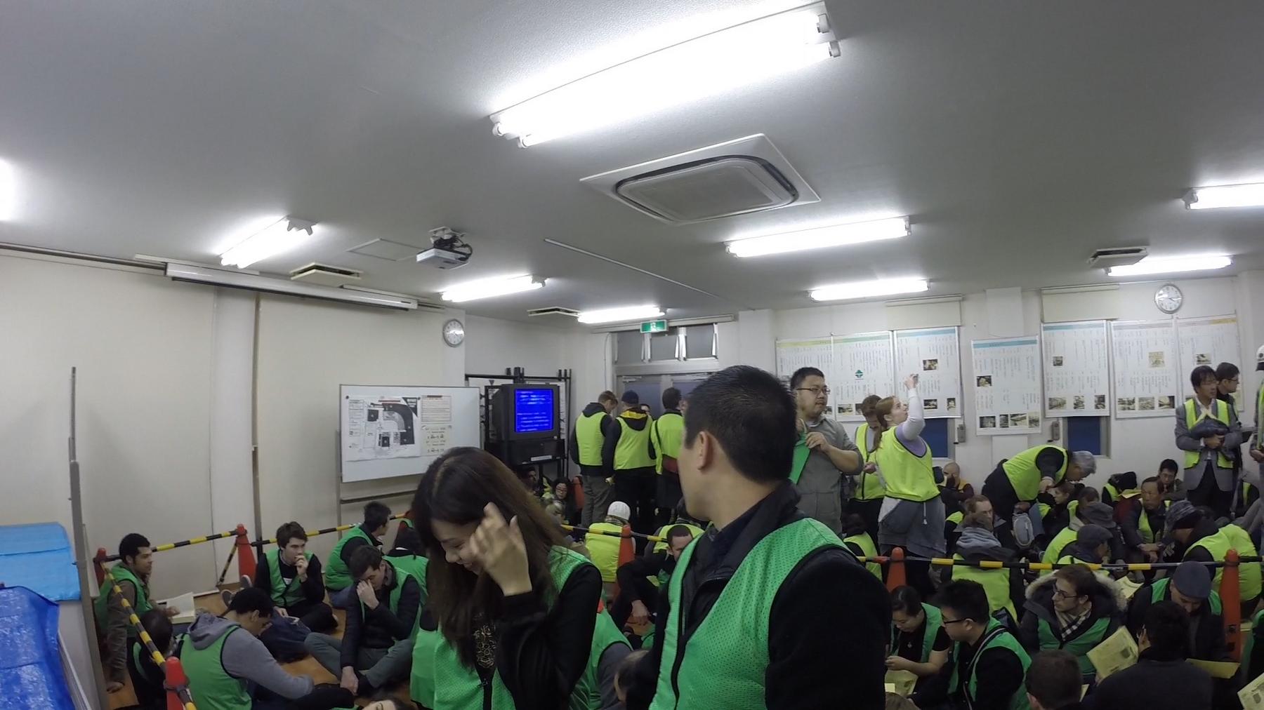 A crowded room of visitors wearing bright green vests, many sitting on the floor, waiting before the Tsukiji tuna auction.