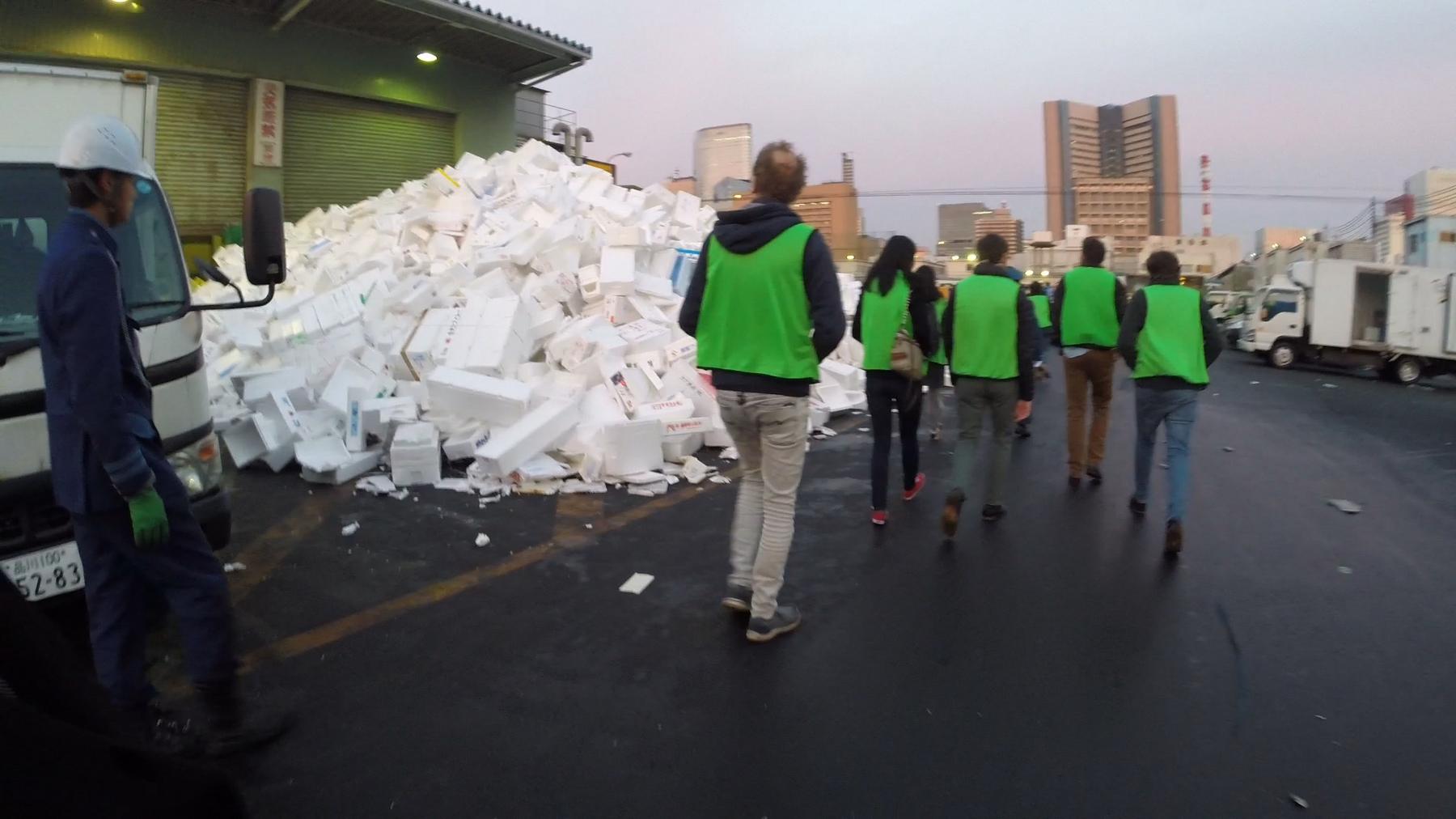 Tourists in green vests walk past a large pile of foam boxes on a wet street at Tsukiji Fish Market.