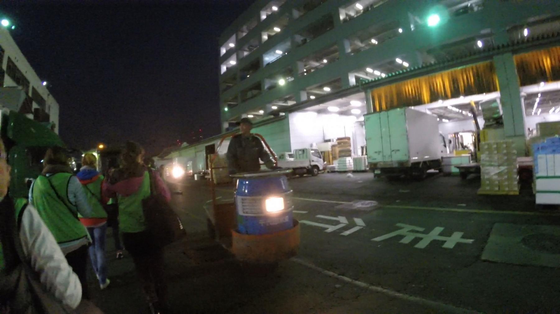 A worker on a motorized cart drives past people in reflective vests and parked trucks at the brightly lit Tsukiji Fish Market in the early morning darkness.