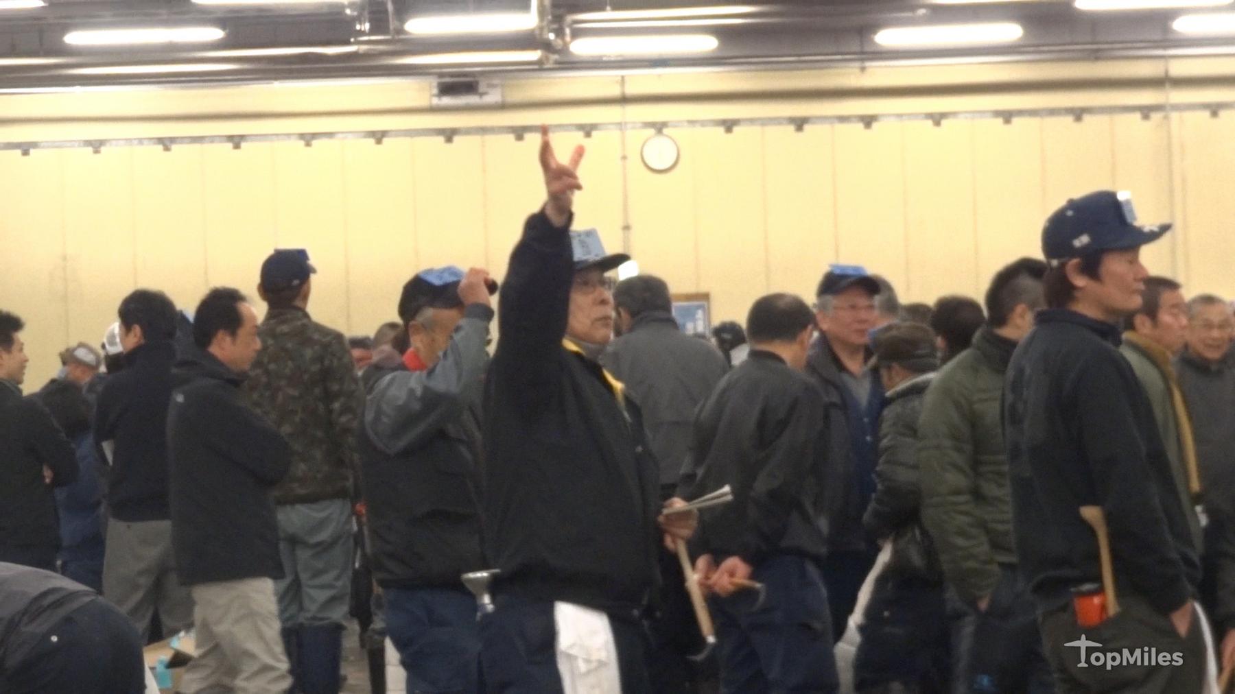 A man raises his hand to bid at the Tsukiji Fish Market tuna auction, surrounded by a crowd of other men.