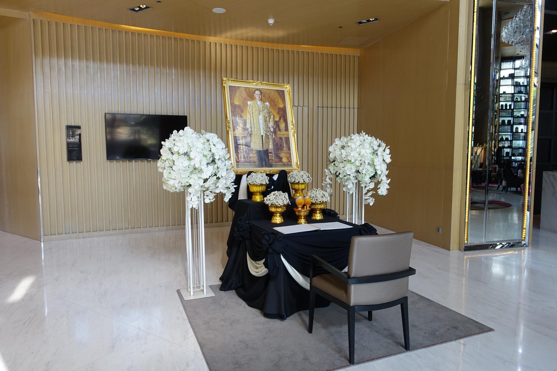 A memorial display in a hotel lobby featuring a large portrait of a Thai King, a black-draped table with offerings, and white floral arrangements.