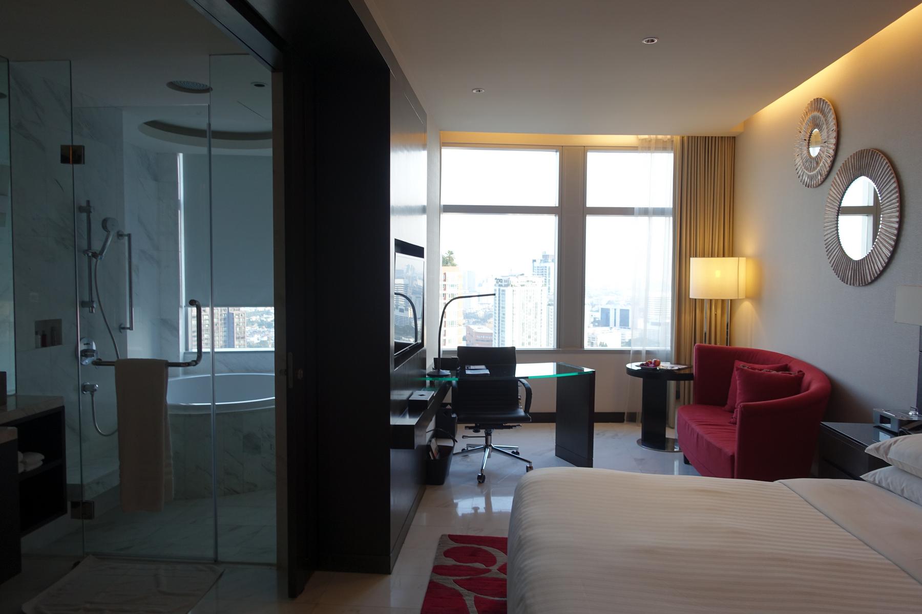 A modern Marriott hotel room featuring a bed, red sofa, desk, and an open-concept bathroom with a bathtub, all with a city view.