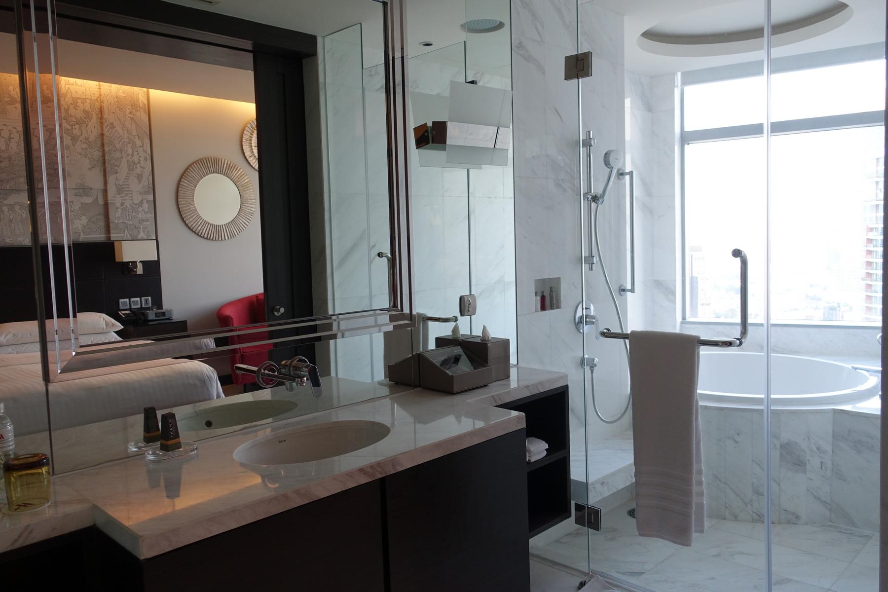 Modern hotel bathroom with marble vanity, glass shower, and tub, reflecting the bedroom.