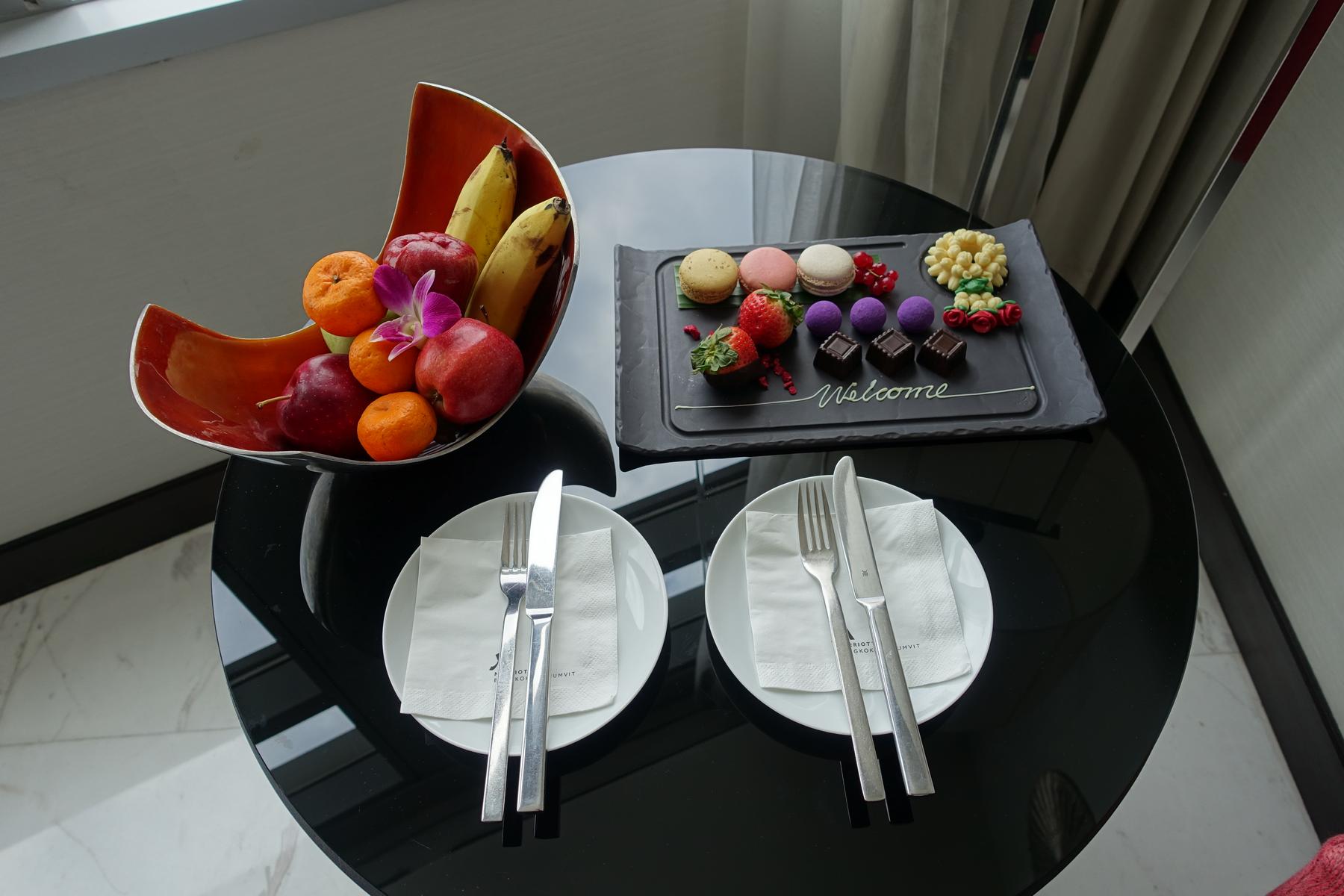 A hotel welcome amenity with a fruit bowl, sweets tray including macarons and chocolates, a Thai garland, and two place settings.