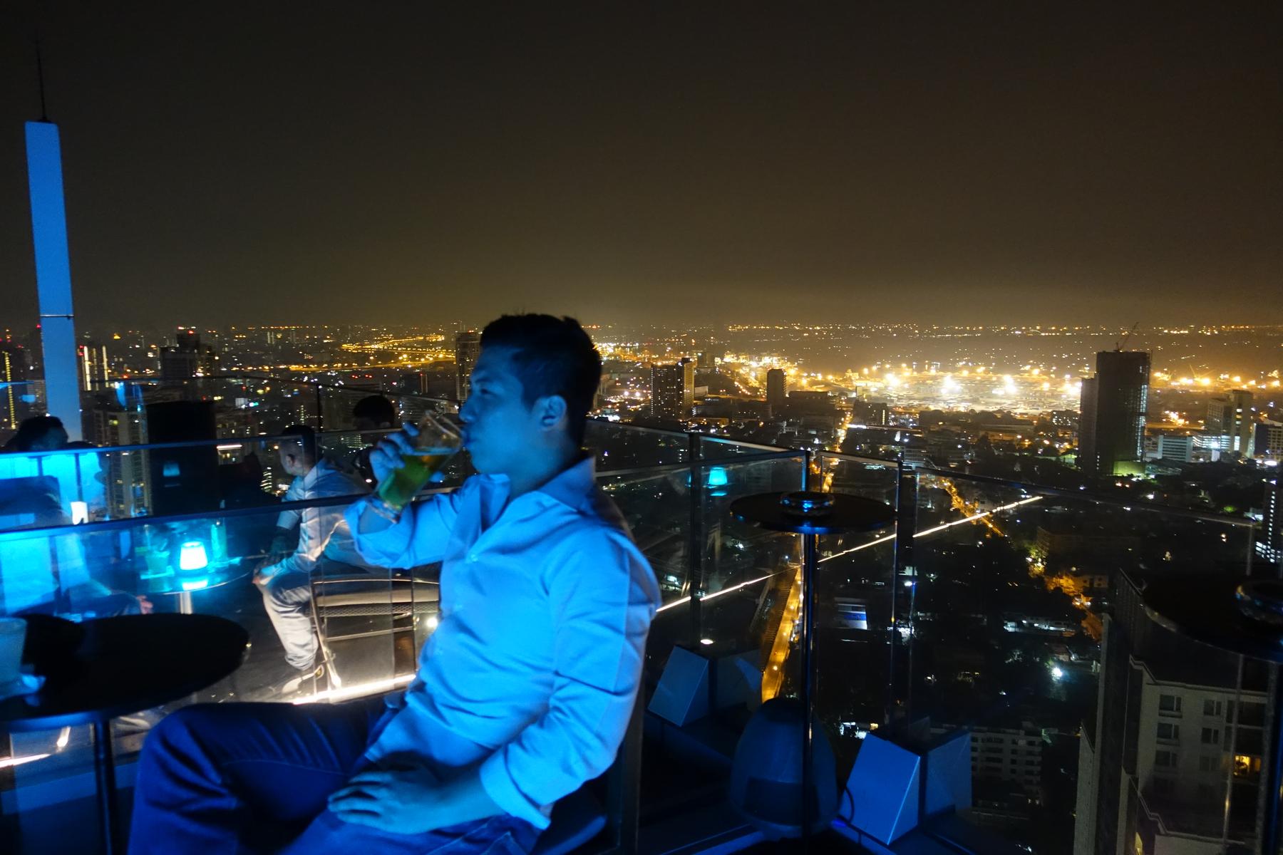A man sips a drink at a rooftop bar overlooking the illuminated Bangkok cityscape at night.