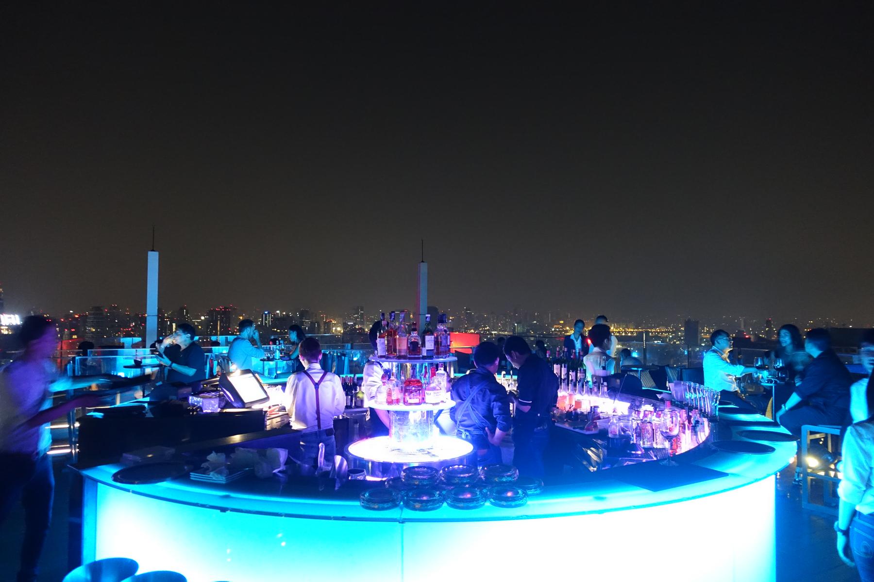 People socialize at a blue-lit rooftop bar overlooking the Bangkok skyline at night.
