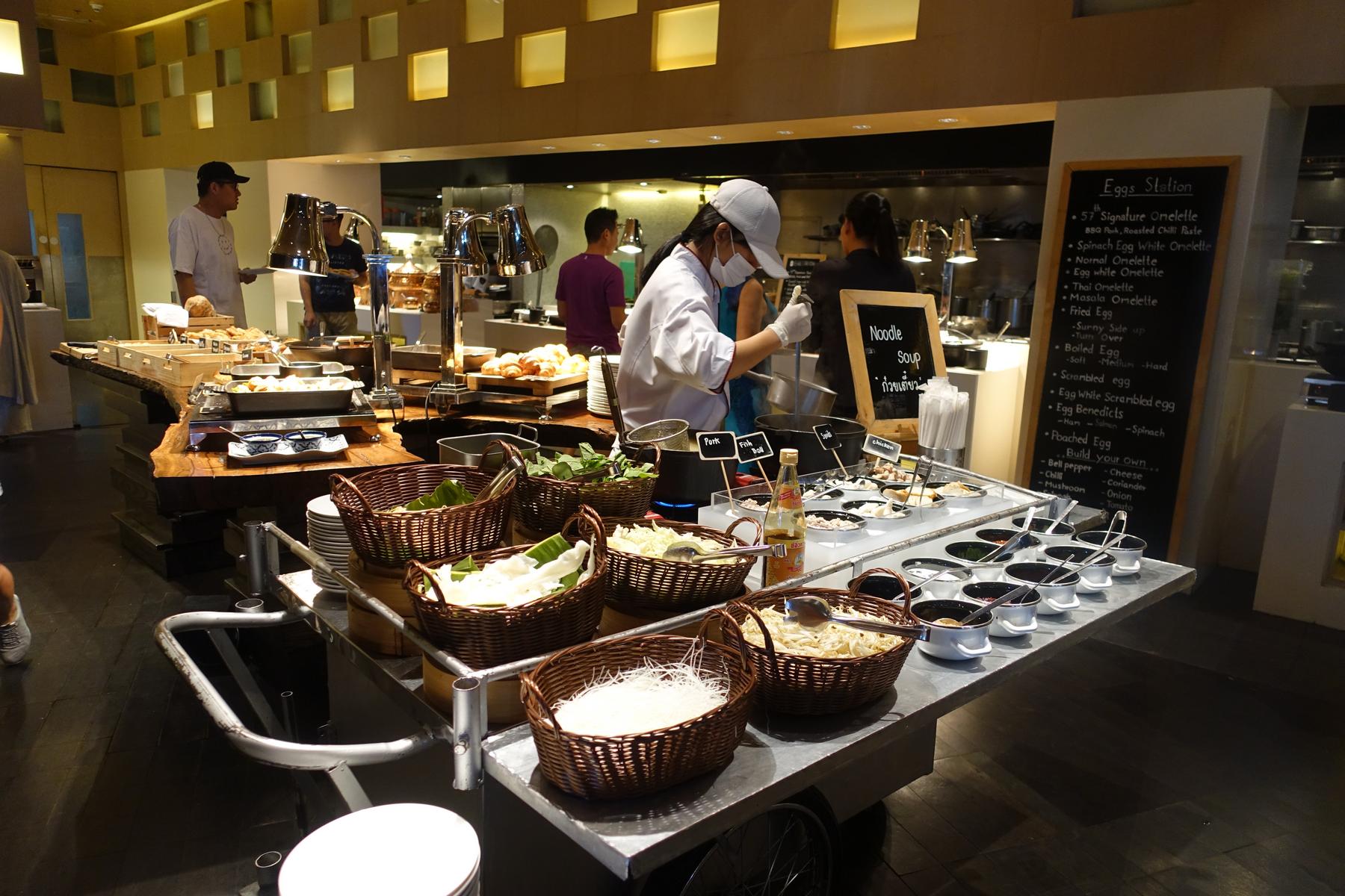 A chef prepares noodle soup at a live cooking station in a hotel buffet, with an Eggs Station blackboard visible.