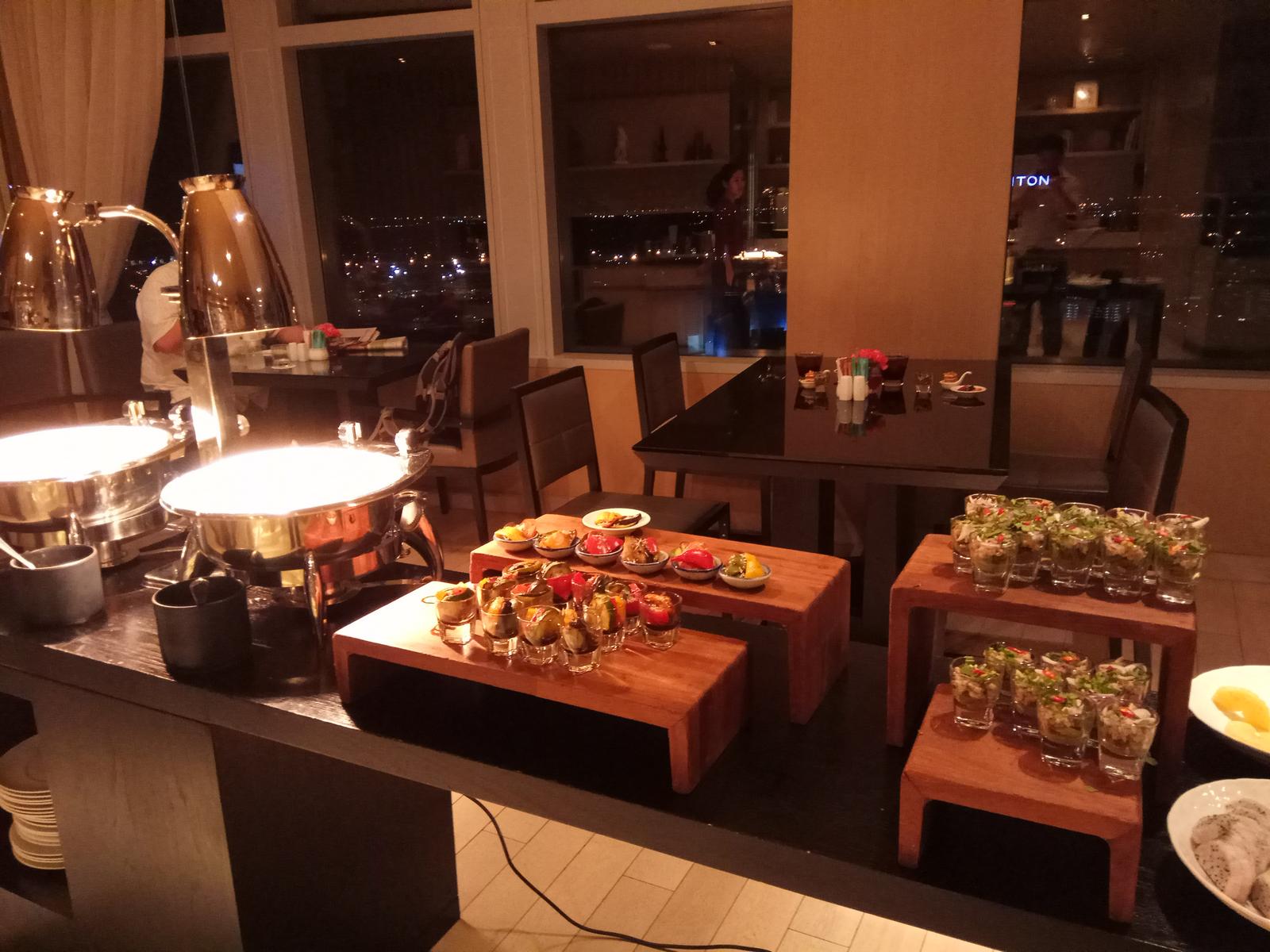 A buffet spread with appetizers, chafing dishes, and a nighttime city view through large restaurant windows.