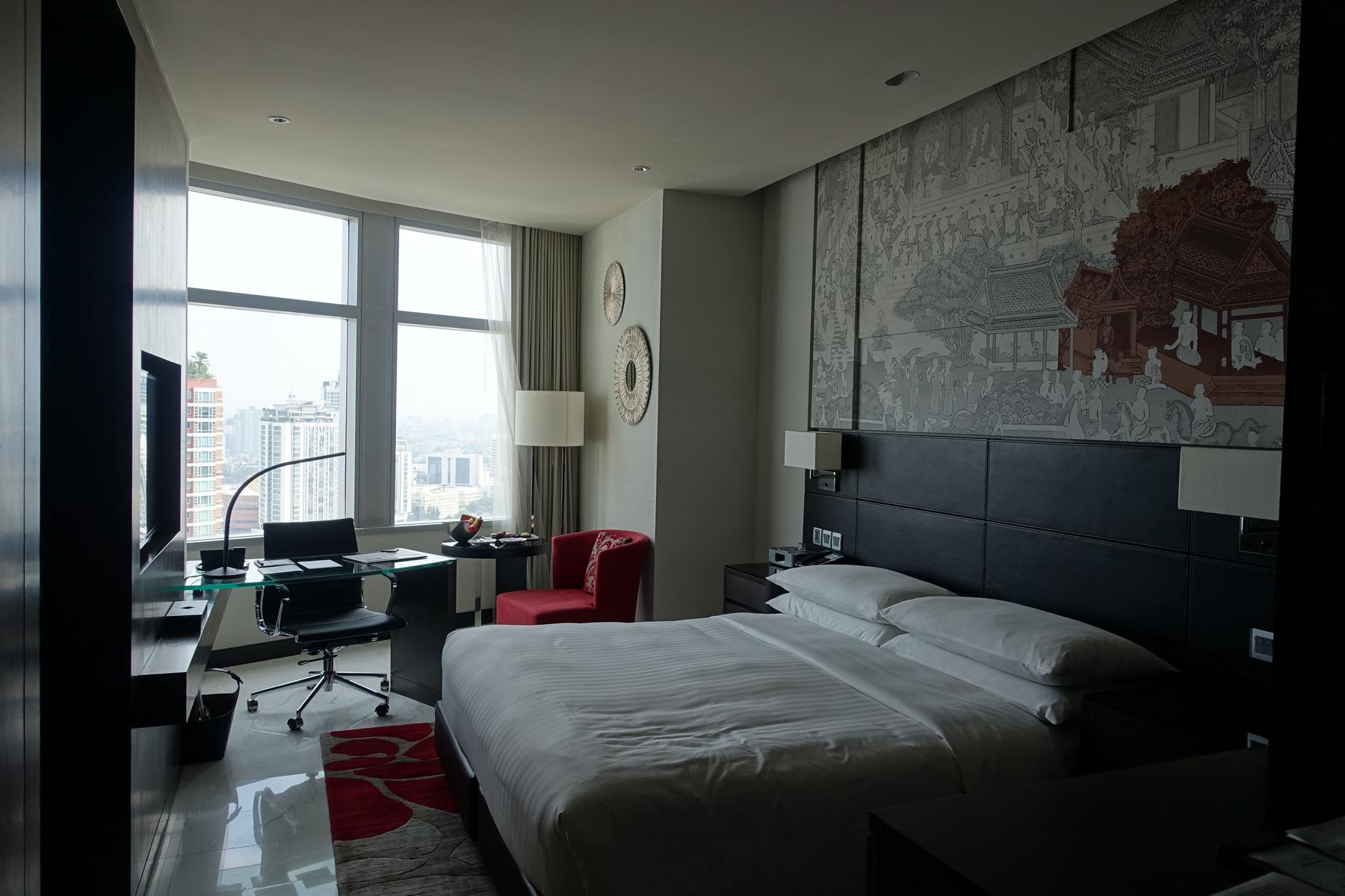 A hotel room with a large bed, a desk by the window with a city view, and a traditional Thai mural.