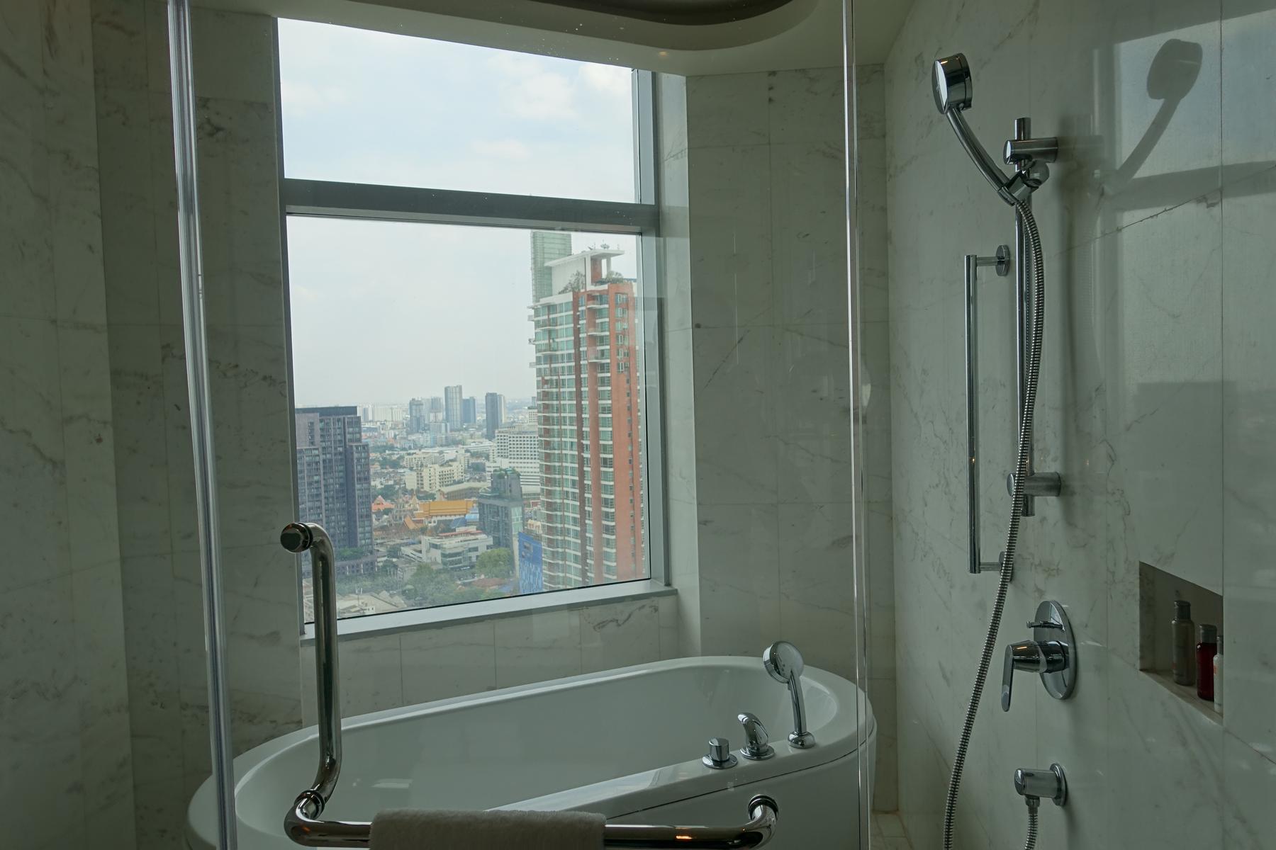 Modern hotel bathroom with a bathtub next to a window overlooking the Bangkok cityscape.