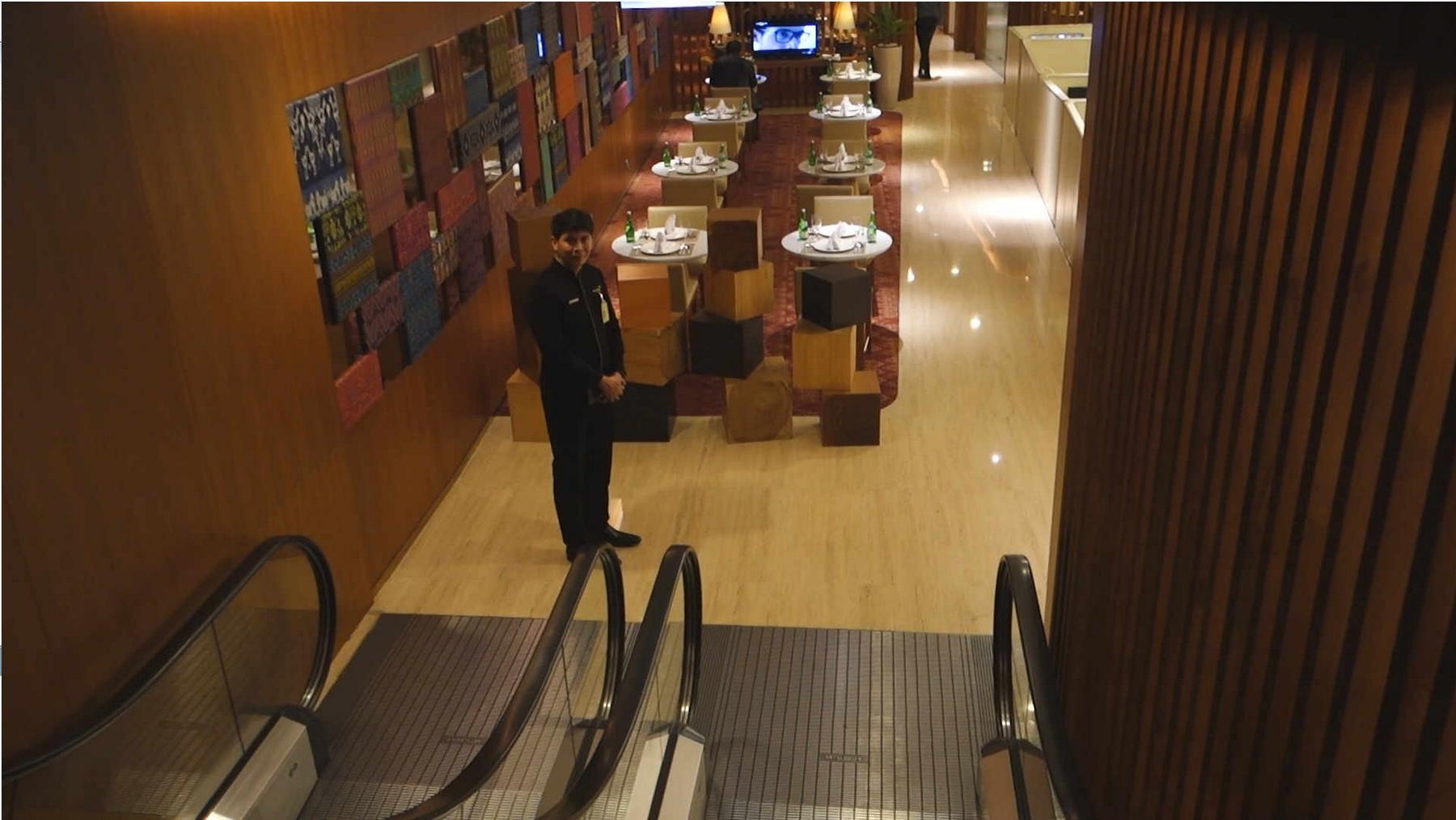 A uniformed staff member stands at the bottom of an escalator in a luxurious lounge with dining tables.