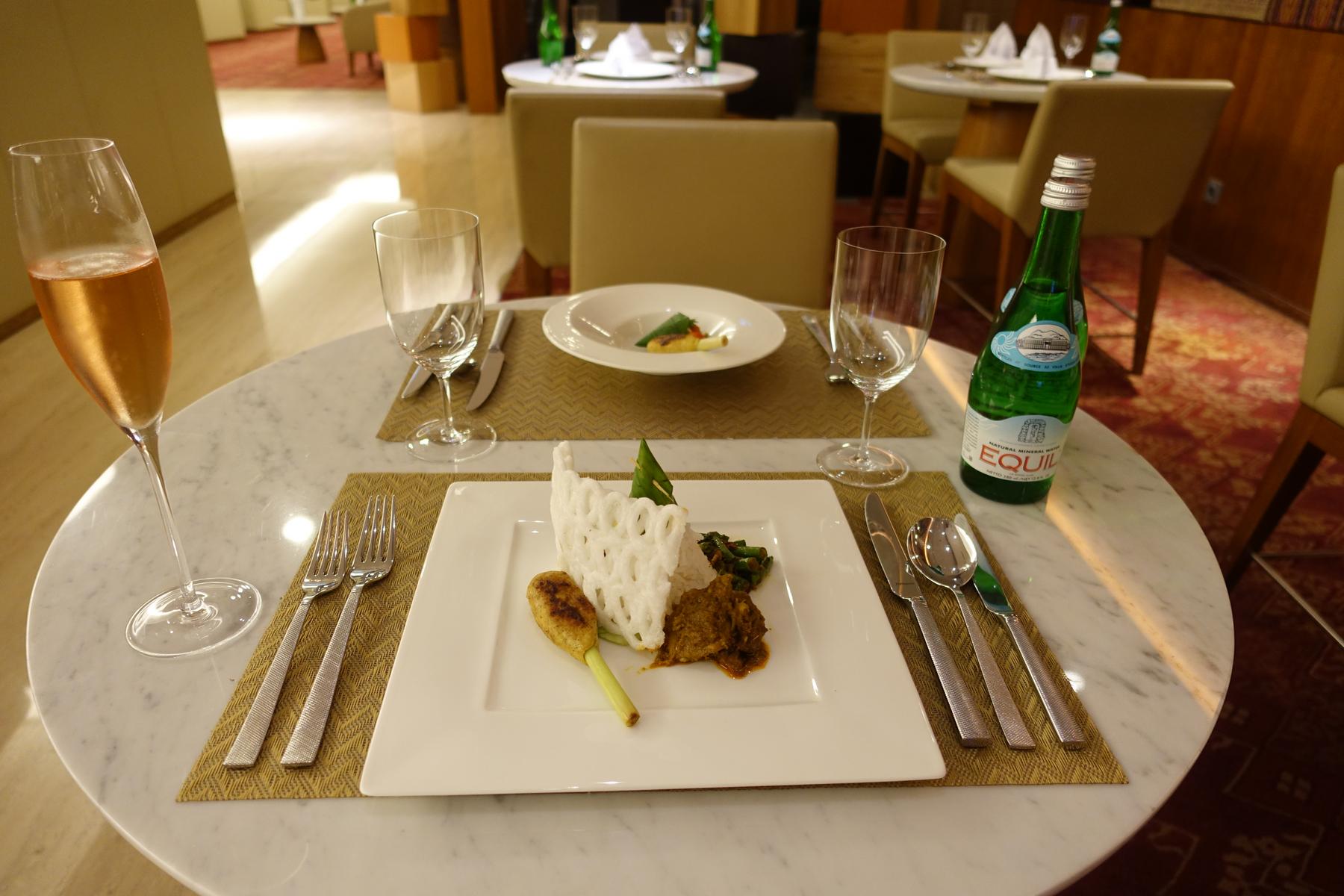 A marble table is set with an Indonesian meal, a glass of rosé champagne, and bottled water in a premium dining lounge.