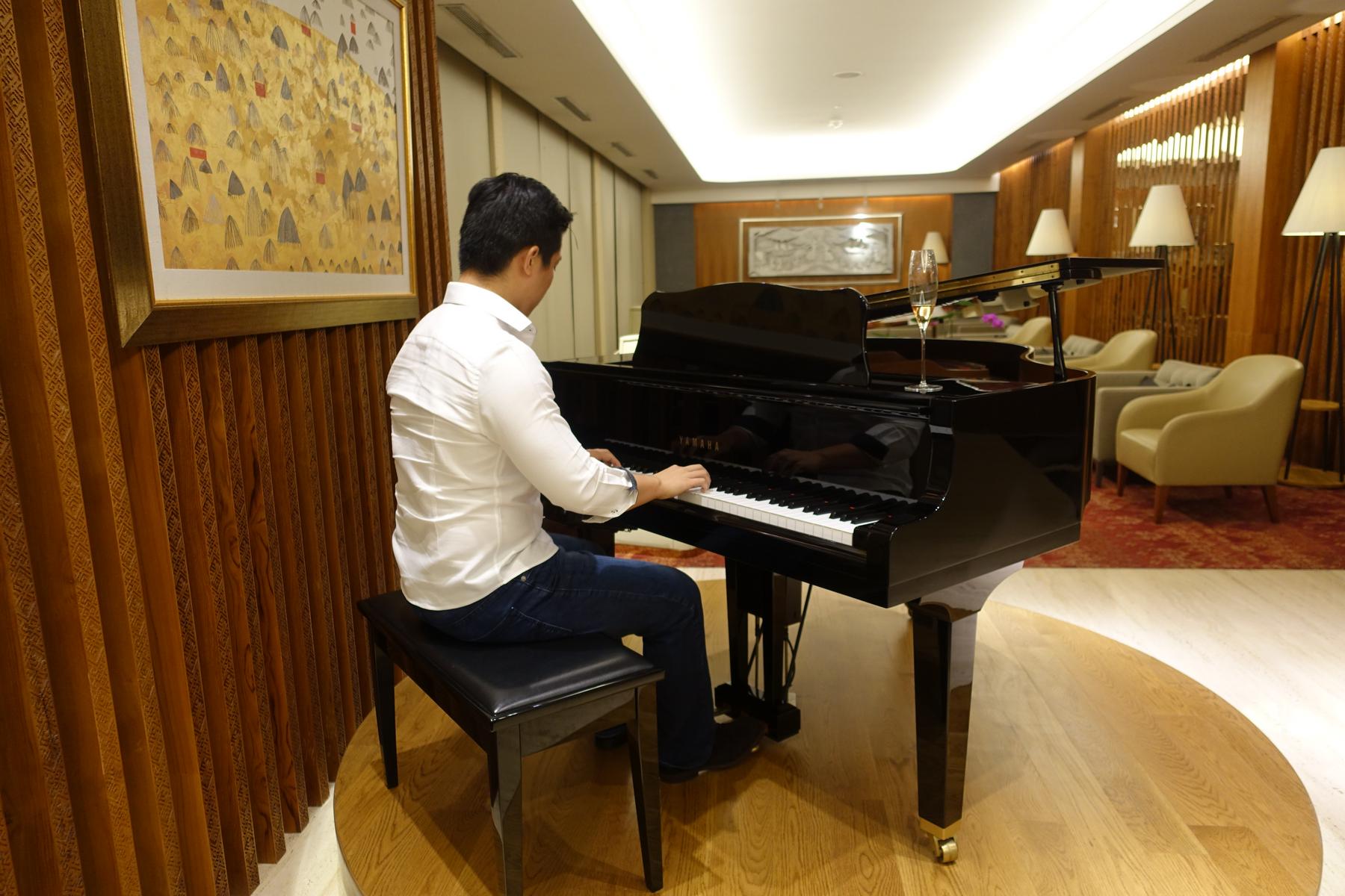 A man in a white shirt plays a black grand piano in a luxurious lounge with a glass of champagne.