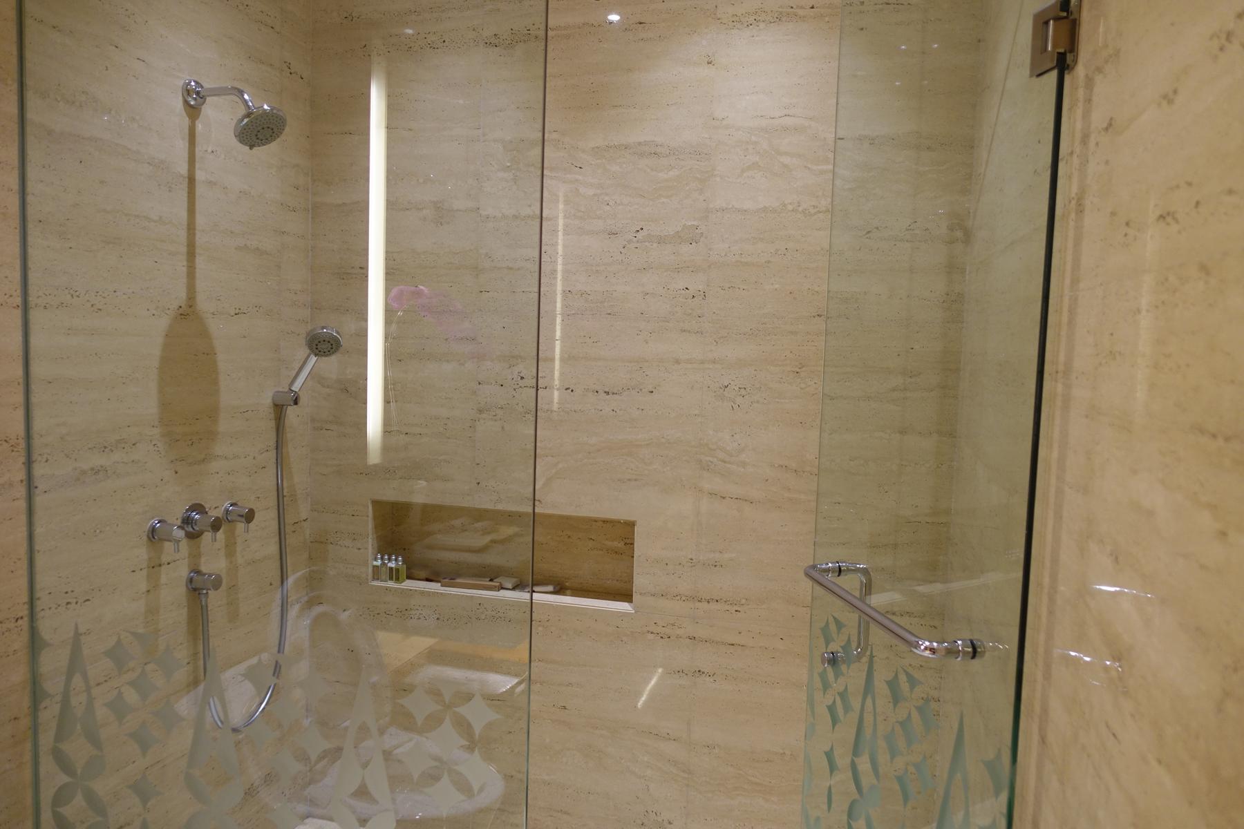 A luxurious shower with light stone walls, glass doors, and both a rain and handheld shower head.