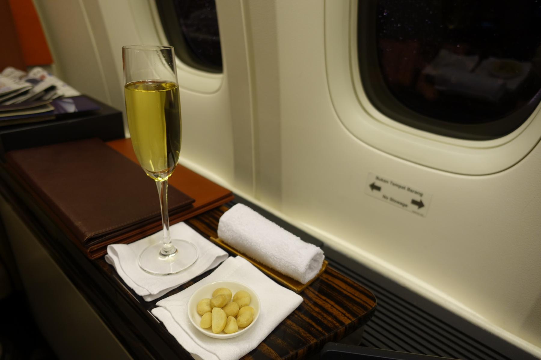 A flute of champagne, a bowl of nuts, and a rolled towel are presented on a wood-grain tray next to an airplane window.
