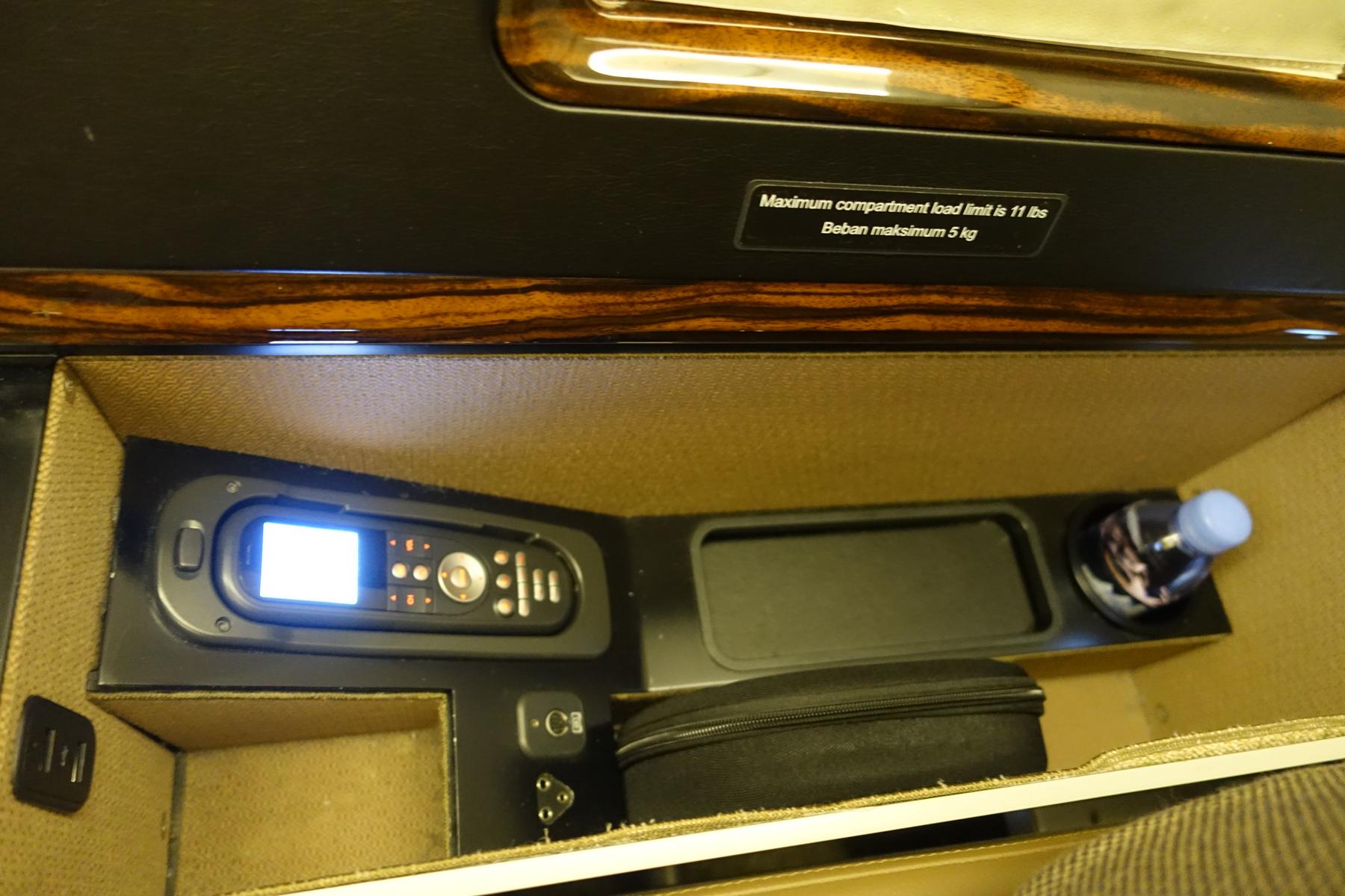 A Garuda Indonesia First Class compartment showing a control panel, power outlet, water bottle, and a load limit sign.