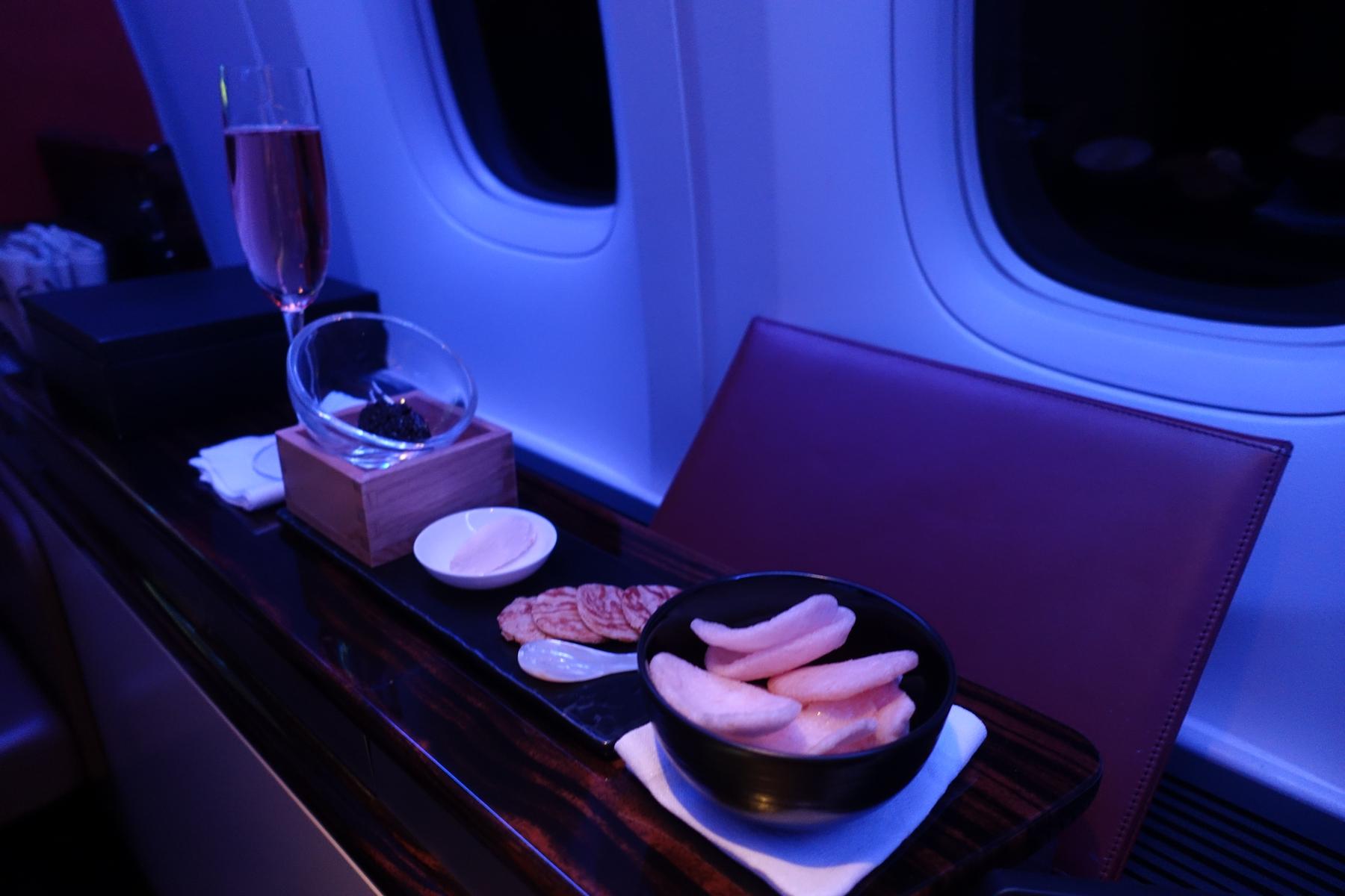 A first-class airplane cabin table with champagne, caviar, sliced meat, and pink crackers under blue mood lighting.