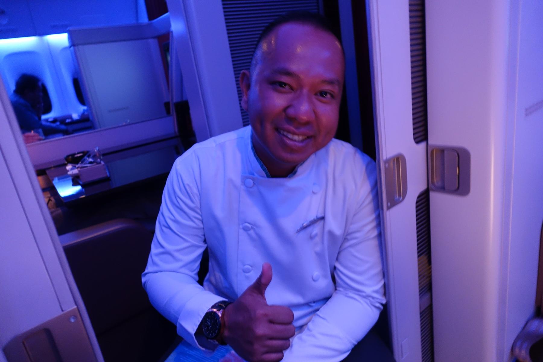 A smiling chef in a white uniform gives a thumbs-up inside a Garuda Indonesia First Class cabin.