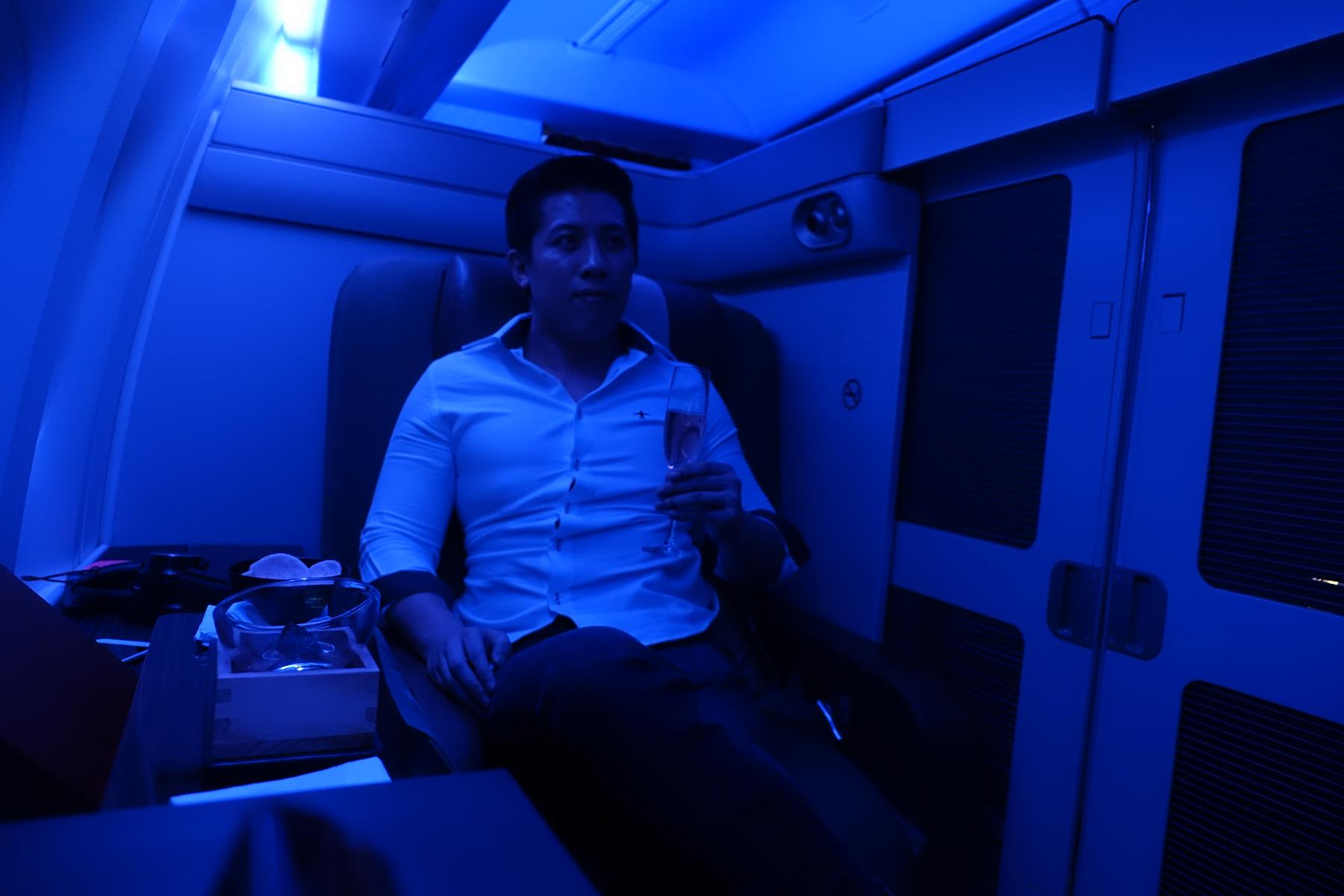 A man holds a champagne flute in a blue-lit first-class airplane cabin.