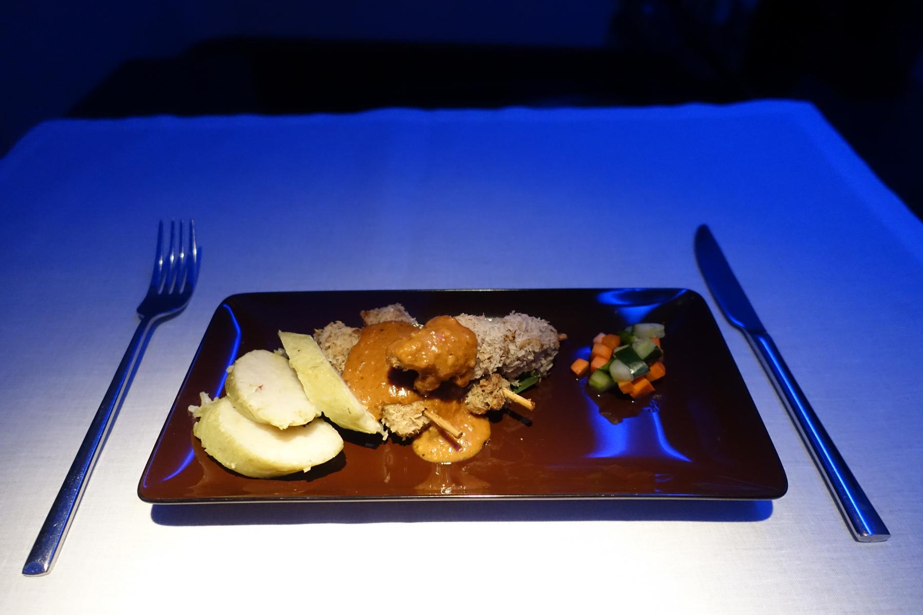 An in-flight First Class meal featuring satay, rice, and vegetables on a brown plate, illuminated by blue cabin lights.