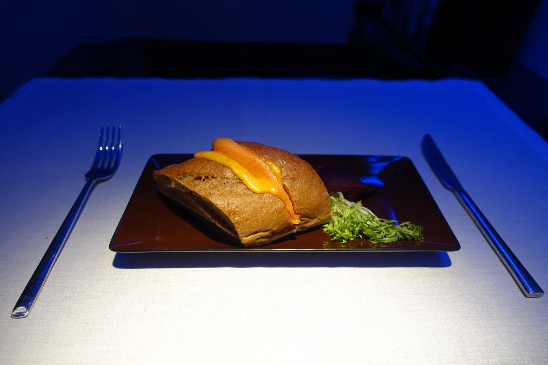 A salmon and cheese sandwich with greens on a brown plate, flanked by a fork and knife, illuminated by blue ambient light.