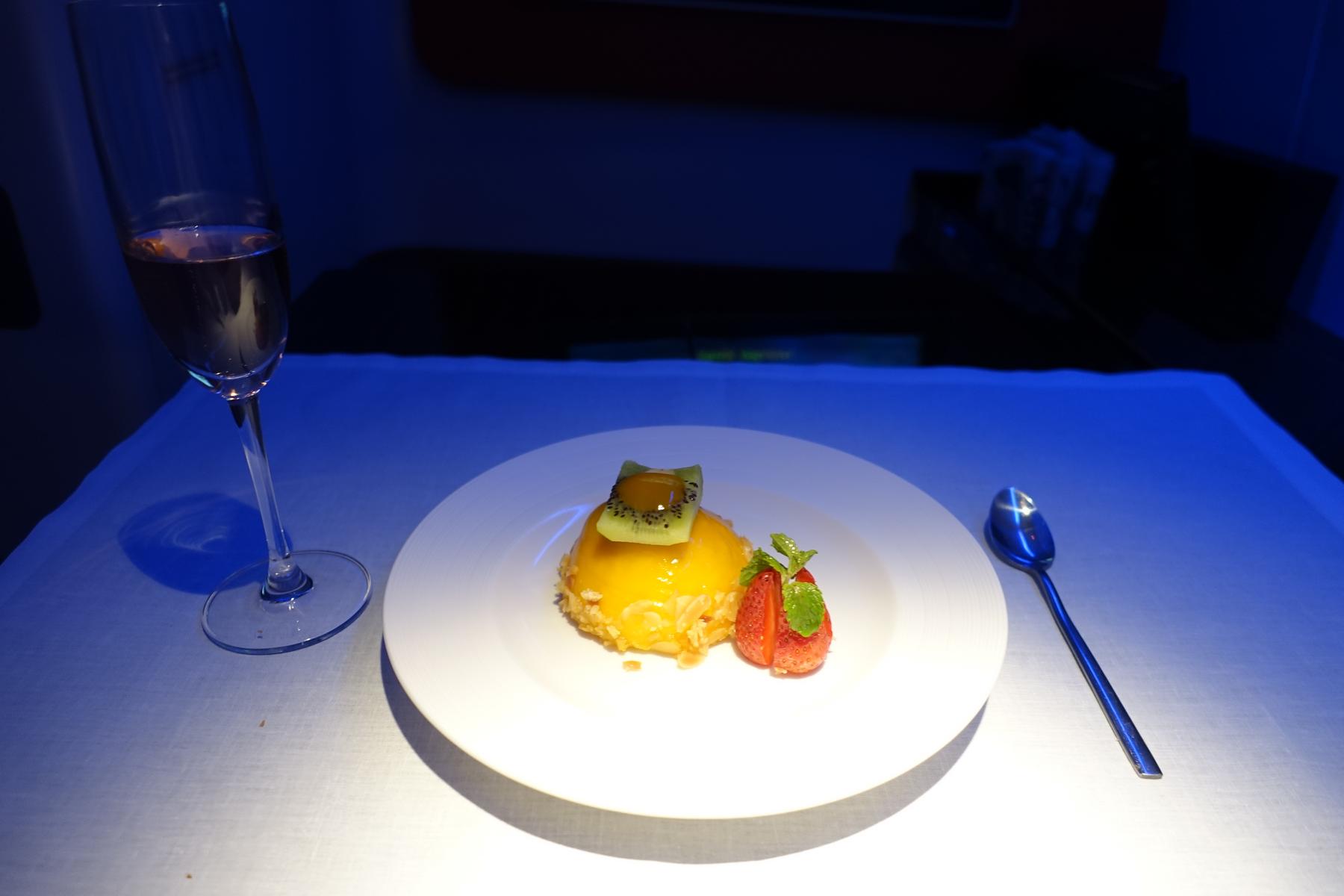 A beautifully plated fruit dessert with kiwi and strawberry is served alongside a glass of champagne on a white table in a first-class cabin.