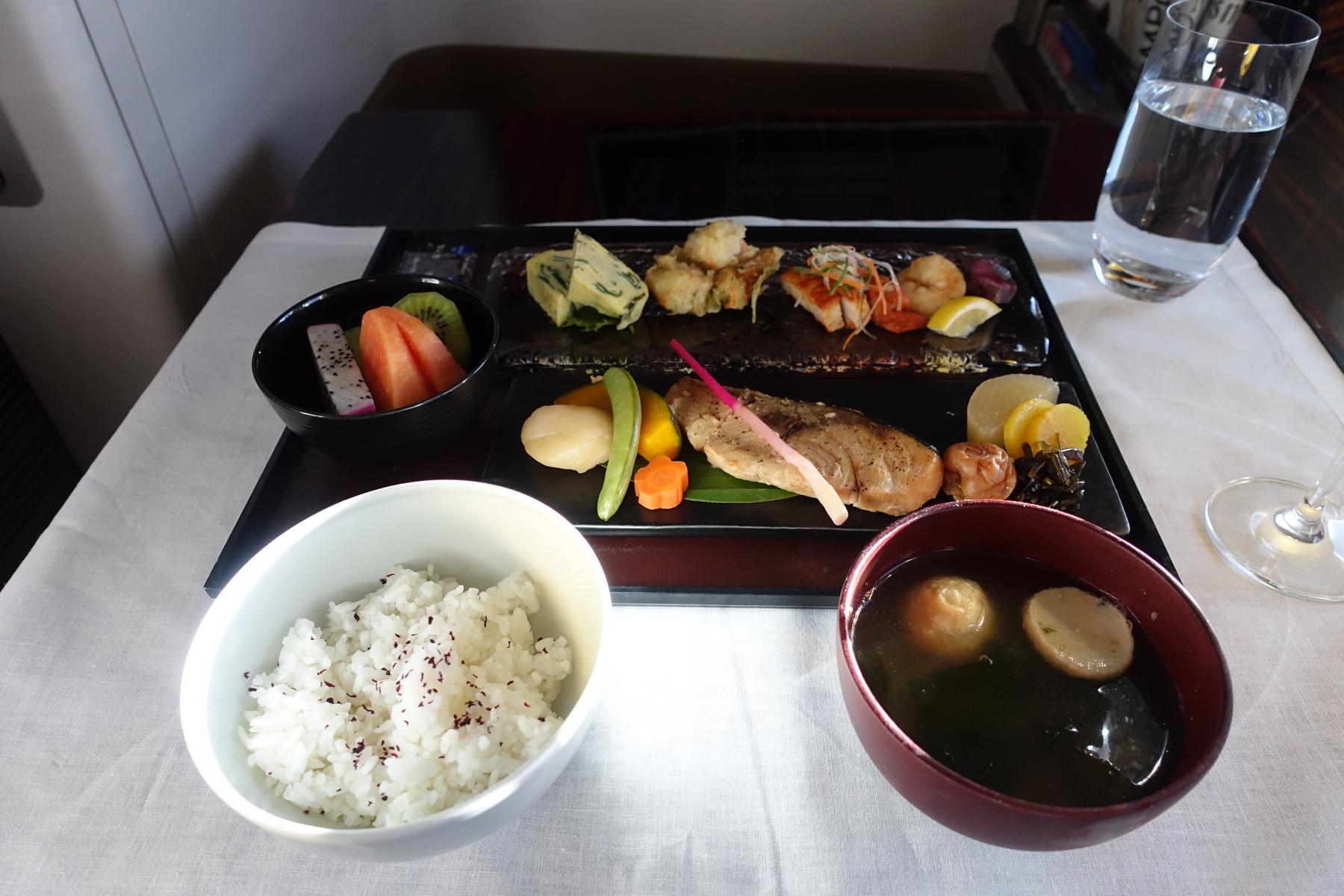 An in-flight First Class Japanese meal featuring grilled fish, rice, soup, fruit, and multiple small dishes.