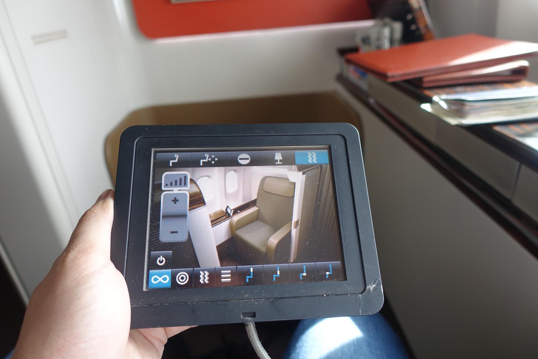 A hand holds a tablet displaying an interactive view of a luxurious airplane seat and cabin controls.