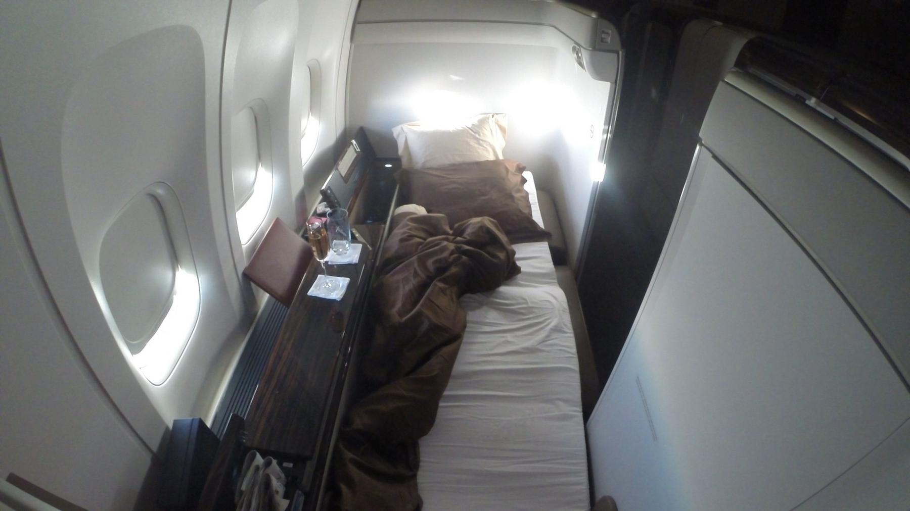 Garuda Indonesia First Class suite with a fully-flat bed, brown duvet, and side table by airplane windows.