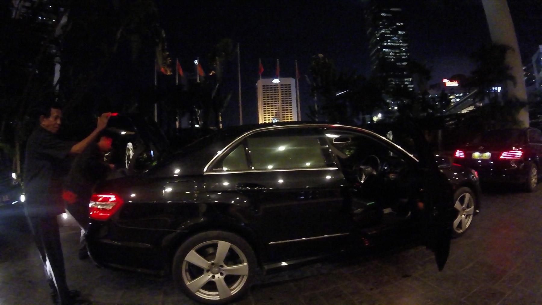A chauffeur loads items into a black luxury sedan at night, with illuminated city buildings in the background.