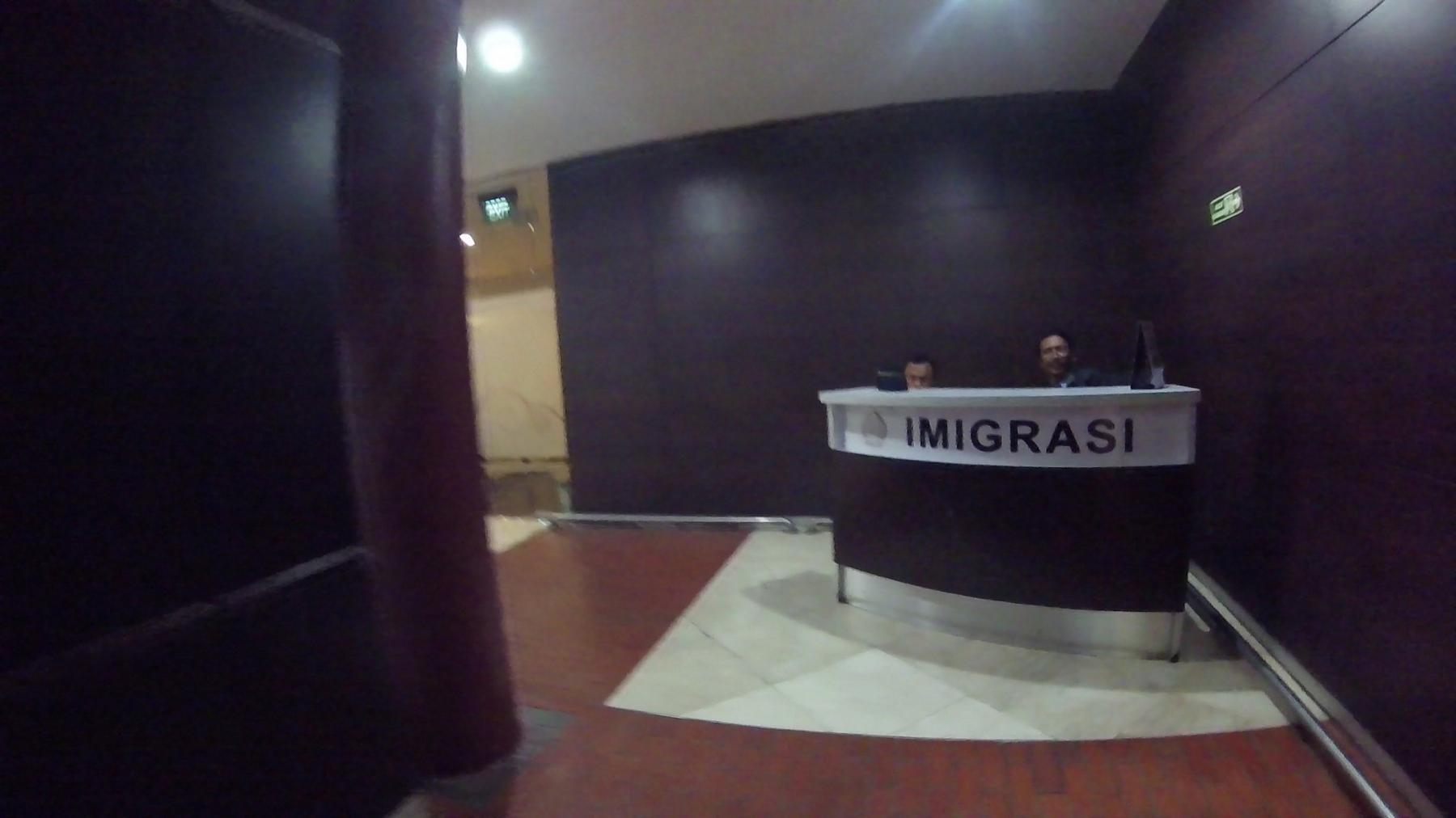 Two immigration officers stand behind a desk labeled "IMIGRASI".