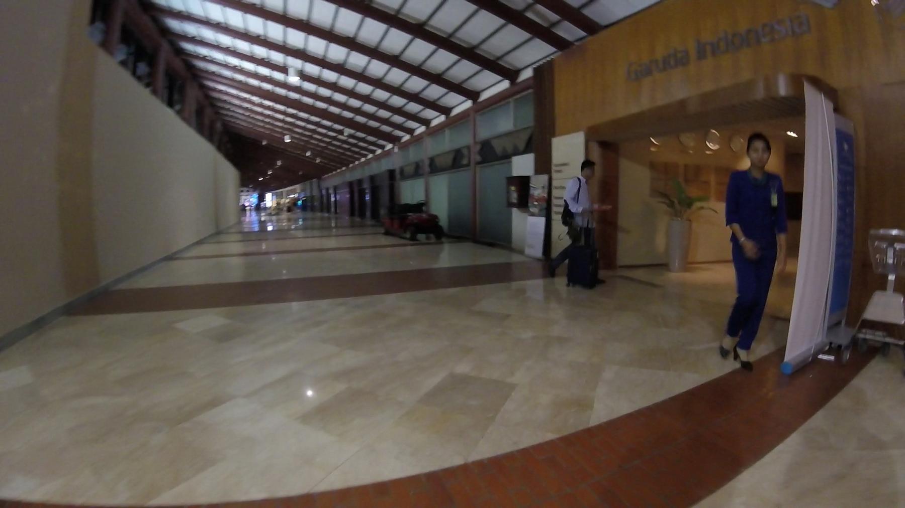 An airport terminal hallway with a Garuda Indonesia sign above an entrance and a uniformed staff member in blue.