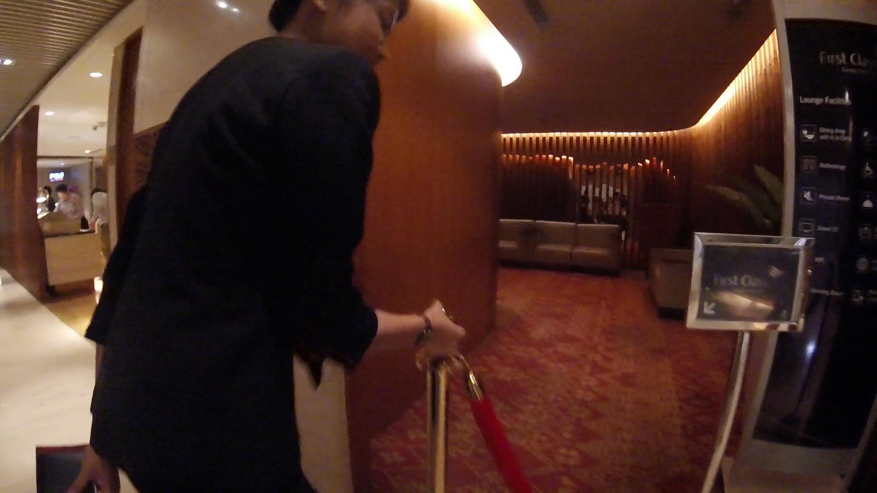 A person pulls back a red rope barrier to enter a First Class lounge.