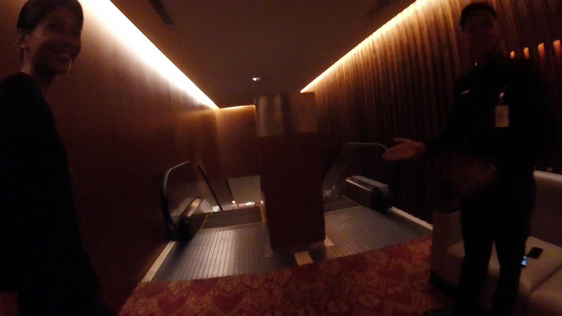 A smiling traveler and a gesturing staff member in a modern, dimly lit airport lounge with warm strip lighting and escalators.