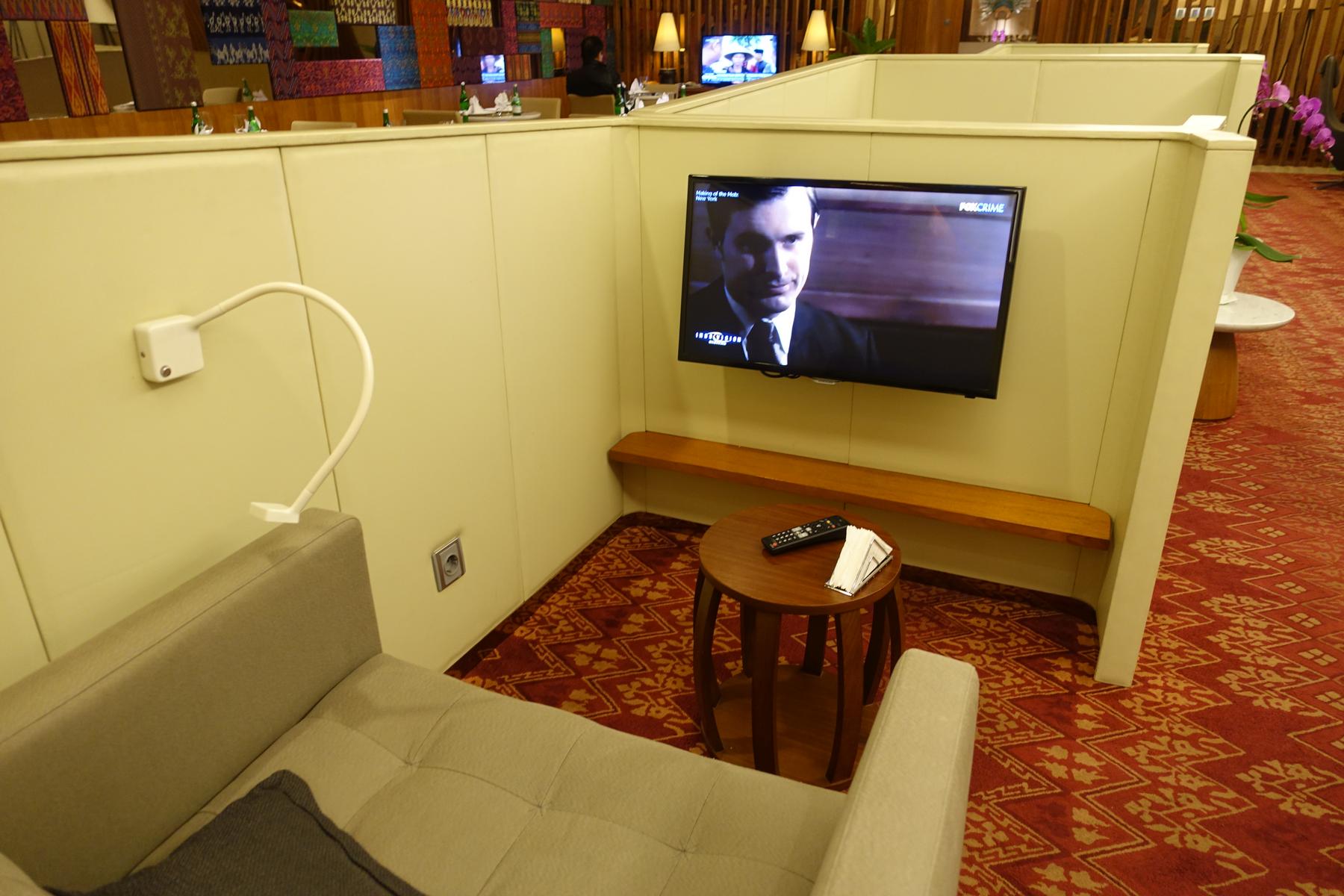 A private lounge seating area with a beige armchair, a TV showing a program, a small wooden table, and a red patterned carpet.