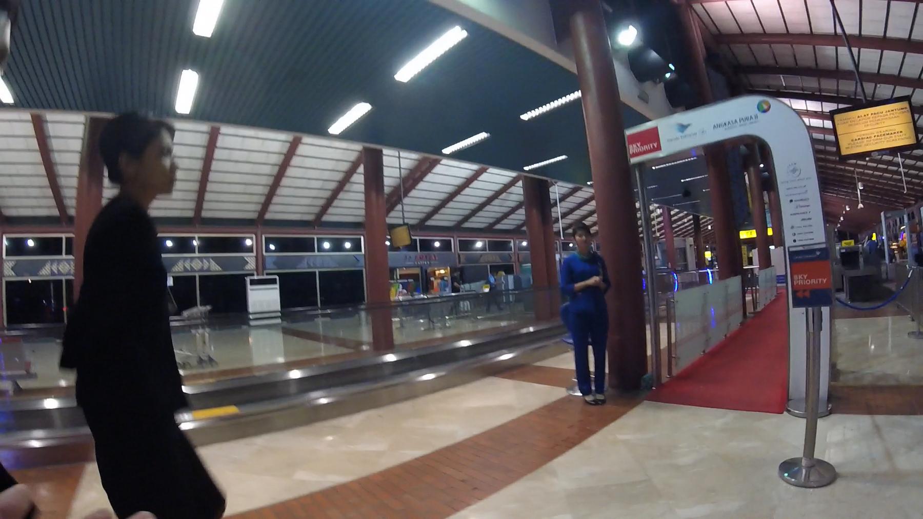 An airport terminal interior showing a Sky Priority archway, an airport employee, and a person walking.