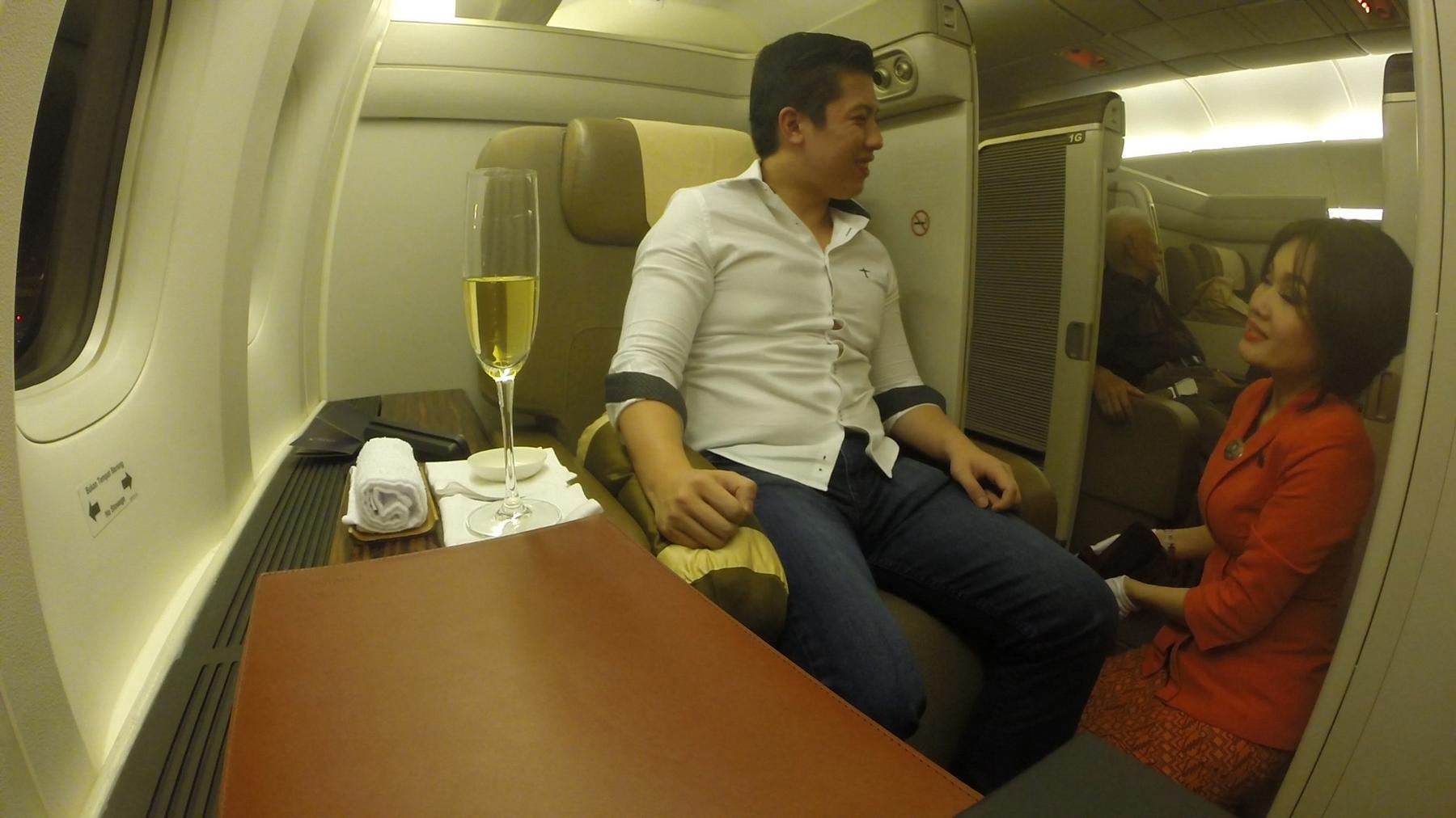 A male passenger in a Garuda Indonesia First Class cabin chats with a kneeling flight attendant, with a glass of champagne on the adjacent table.