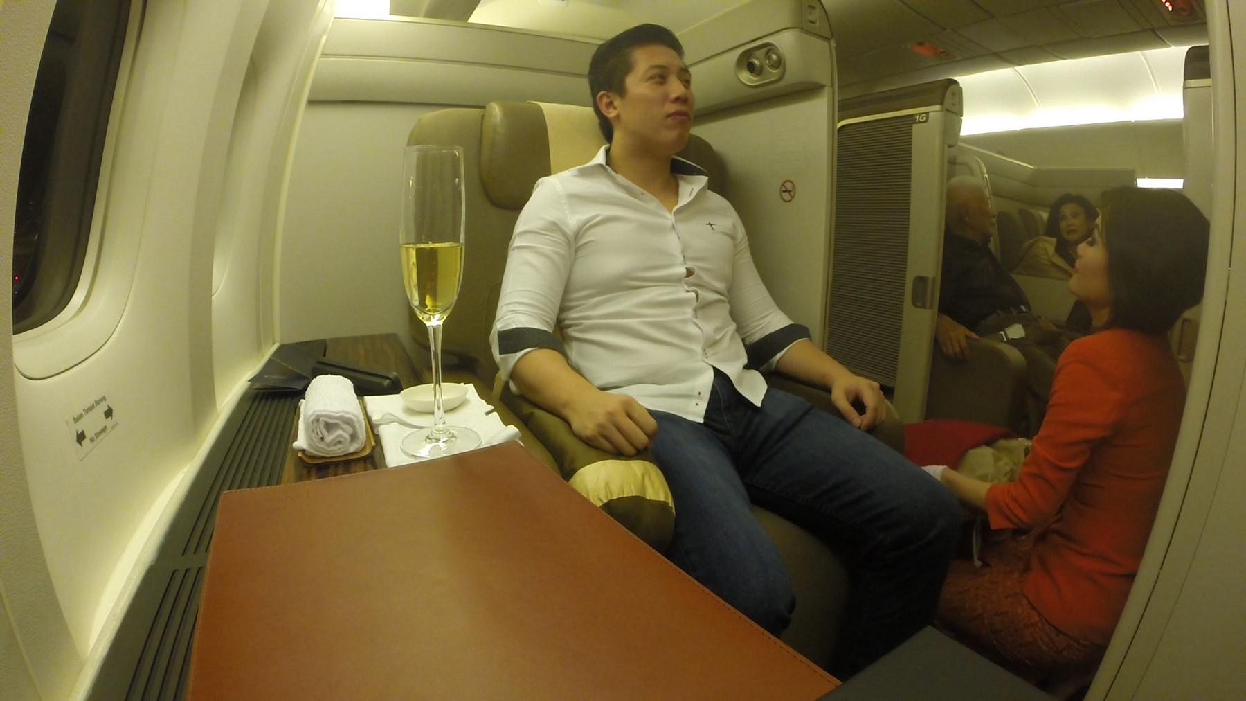 A man sits in a spacious Garuda Indonesia First Class airplane suite with a glass of champagne.