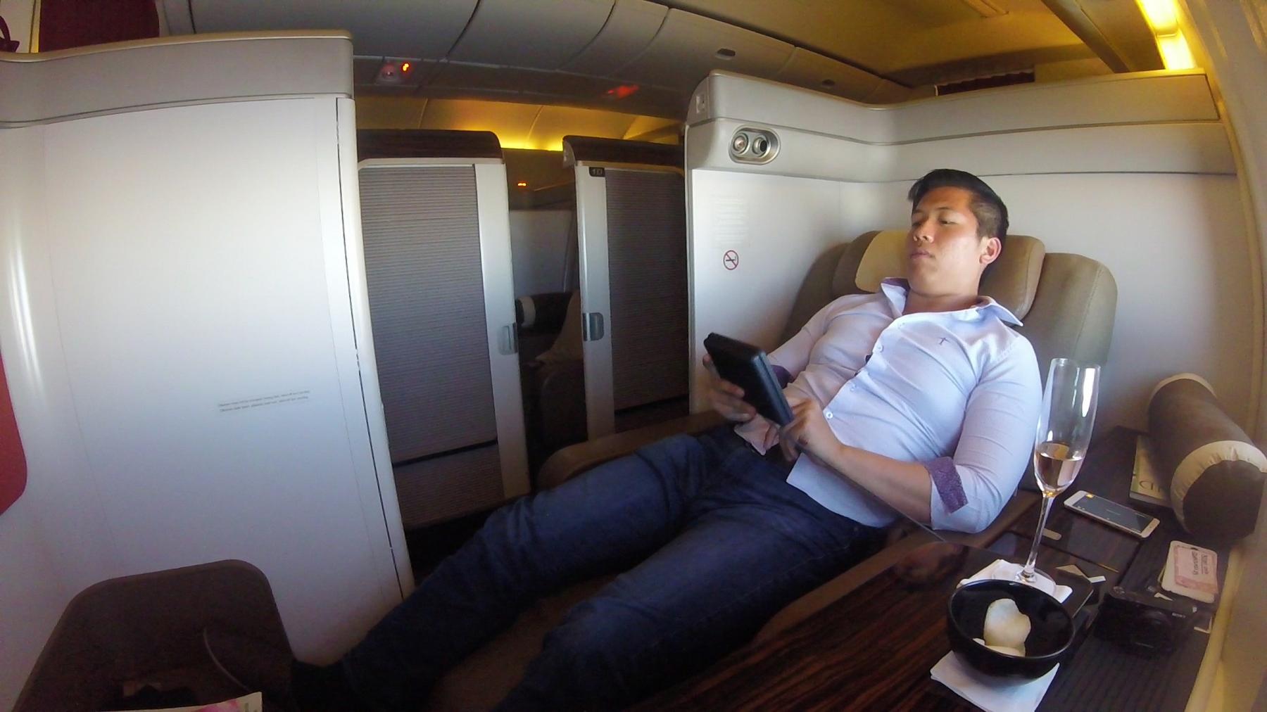 A man lounges in a private first-class airplane suite, holding a tablet with a glass of champagne and snacks on the table.
