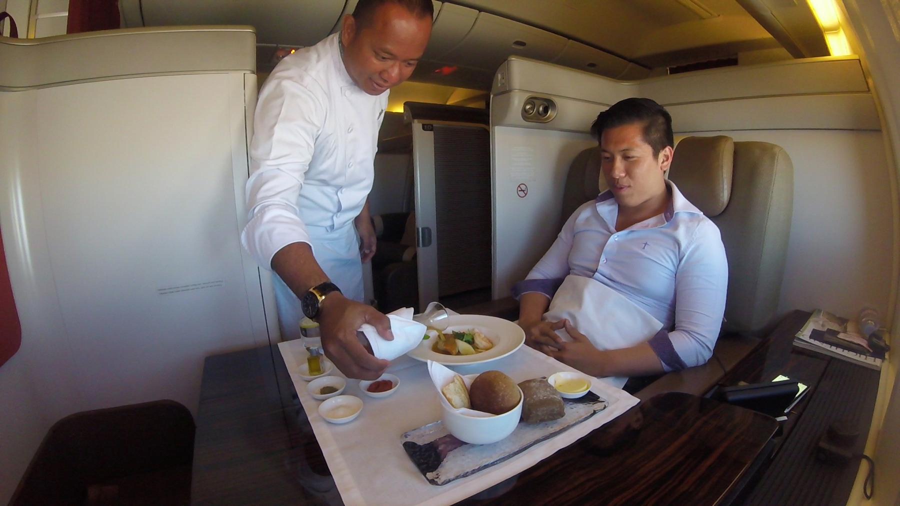 A flight attendant serves an elaborate meal to a passenger in a spacious first-class airplane cabin.