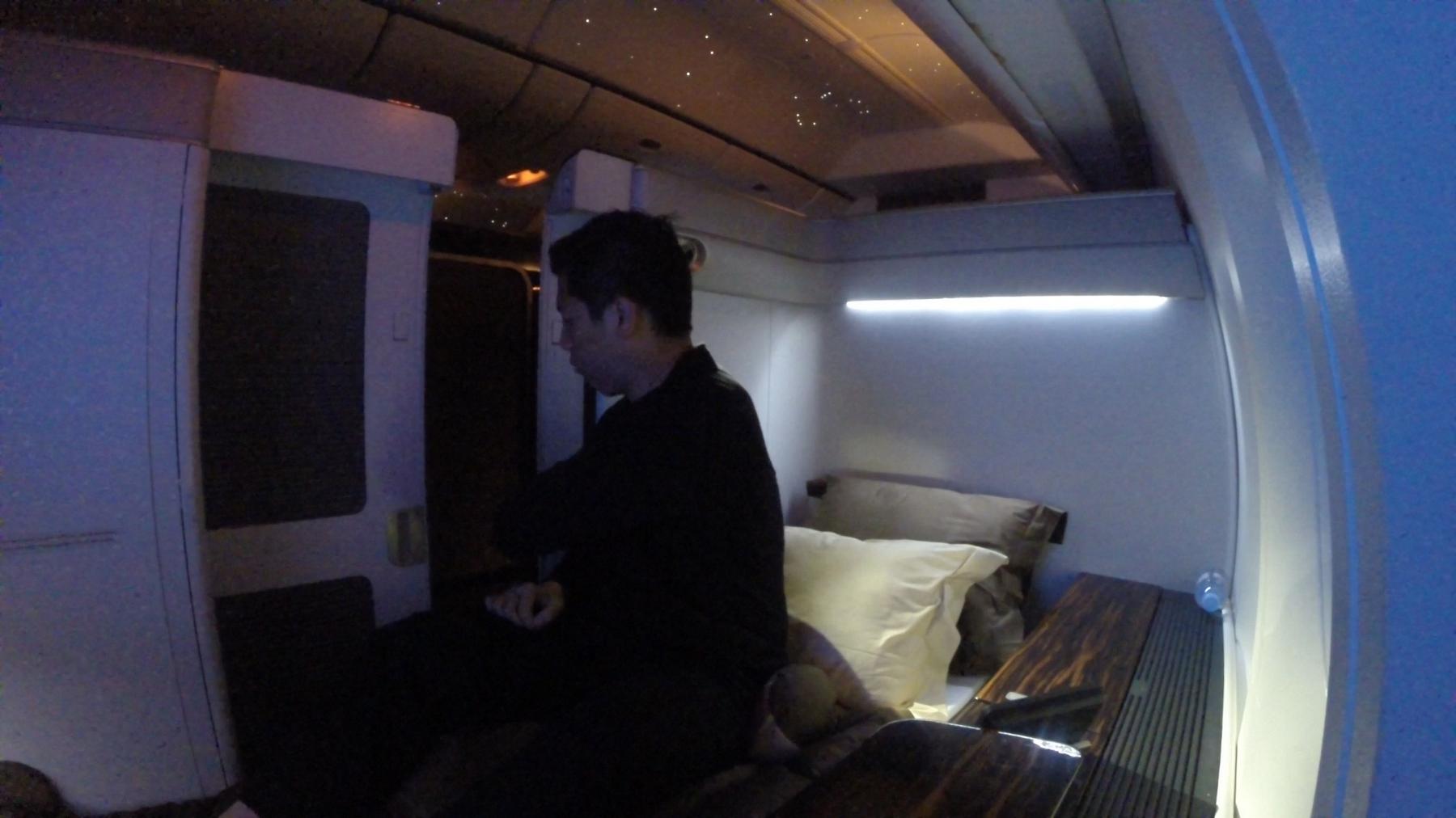 A man sits in a dimly lit first-class airplane suite with a bed and starlit ceiling.