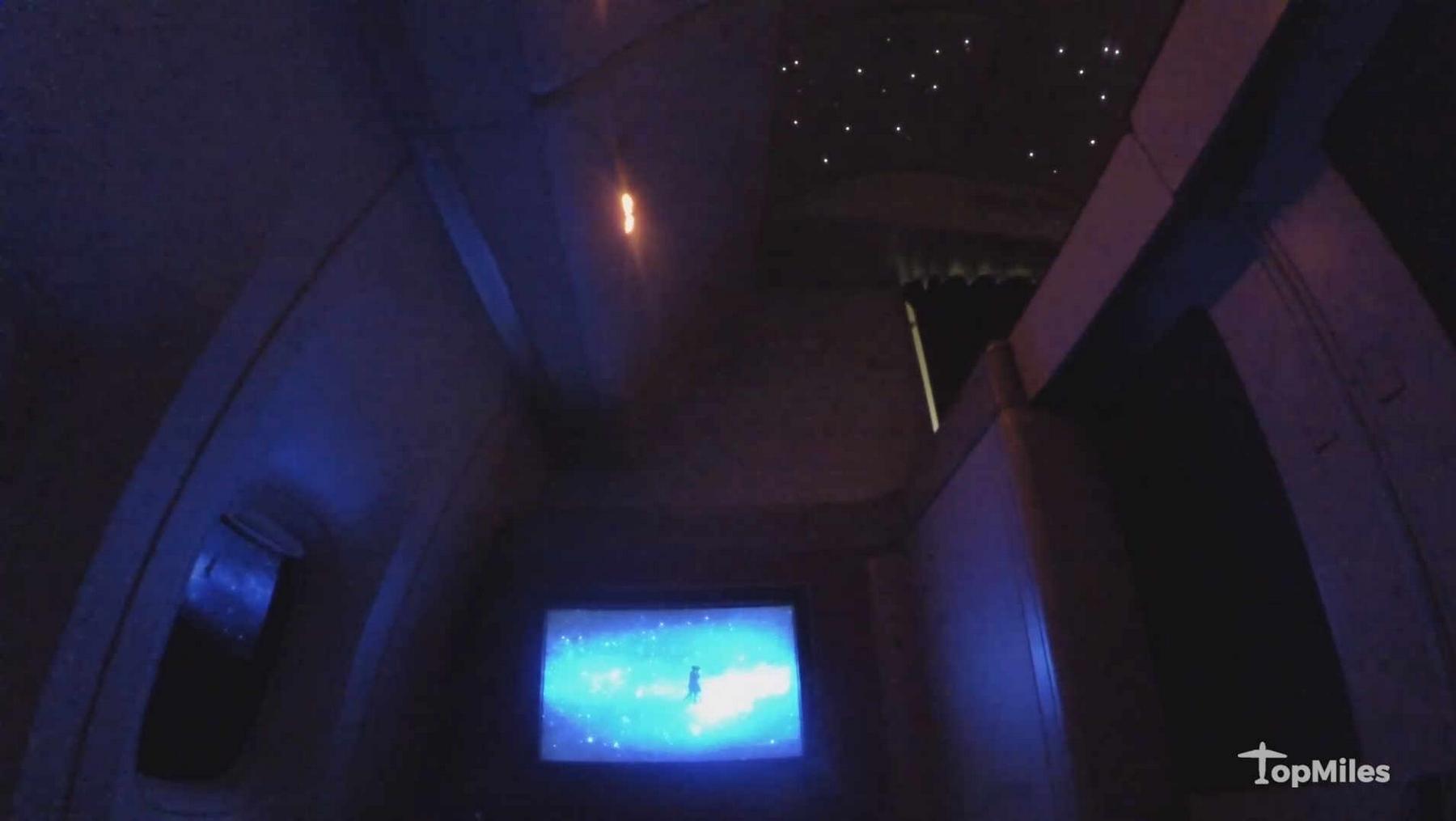 A dimly lit luxury airplane cabin features a starry ceiling and a large screen displaying a vibrant blue cosmic scene.