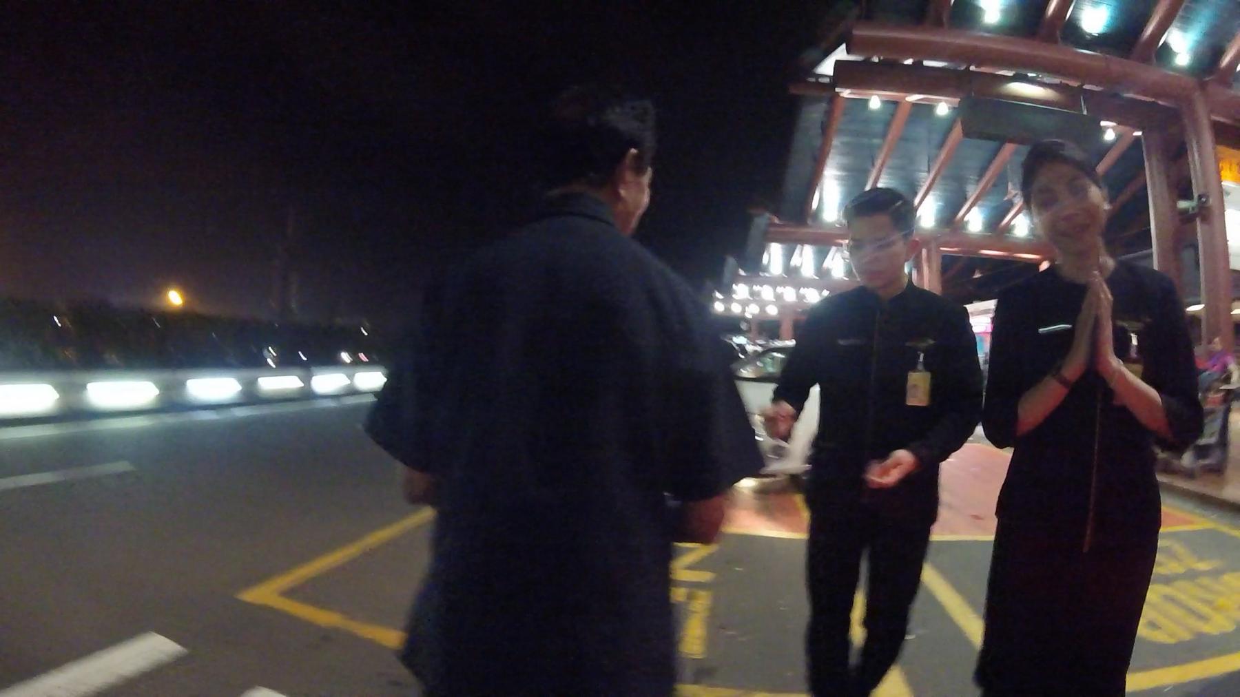 Two Garuda Indonesia staff members, one with hands pressed in a greeting, welcome a passenger at the airport at night.