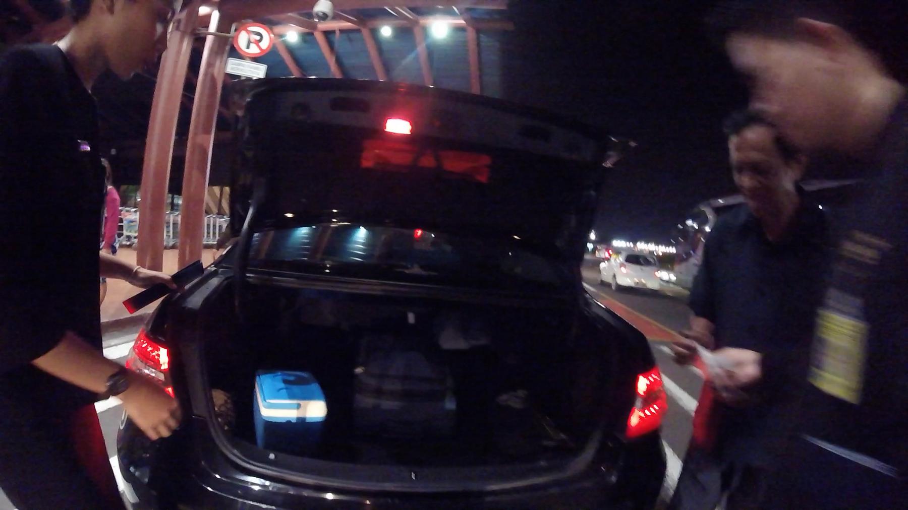 A dark car with an open trunk containing a cooler and a bag stands at night with people around it.