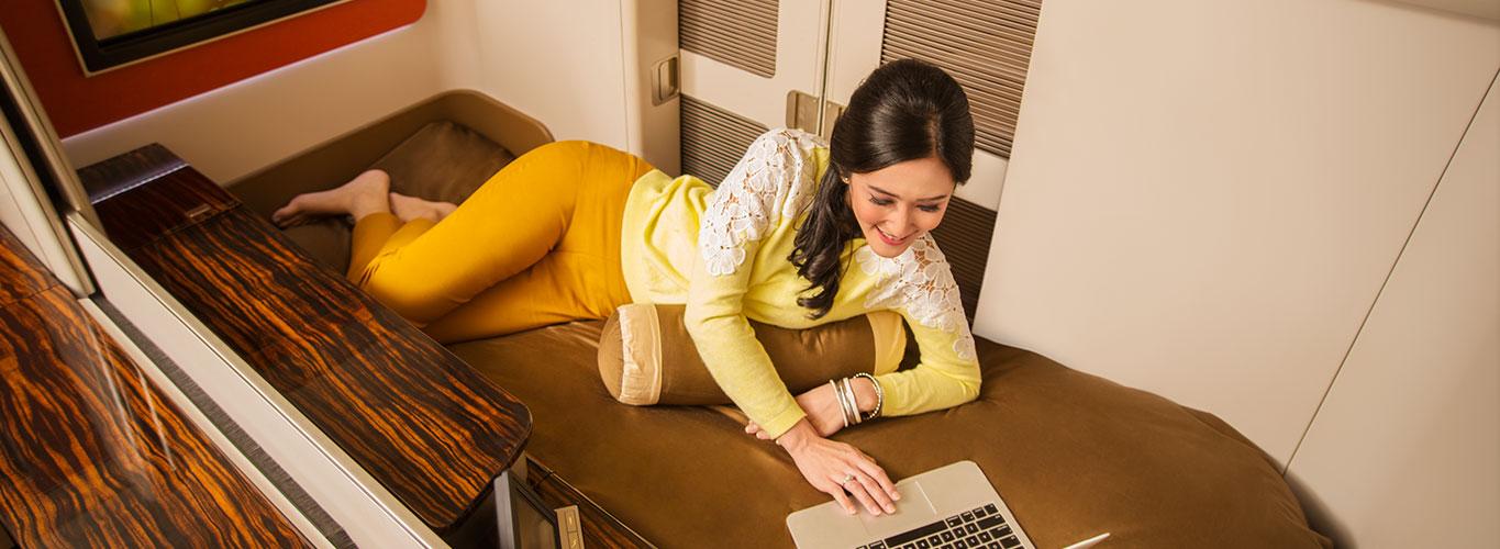 A woman smiles while lying comfortably in a luxurious airplane suite and using a laptop.