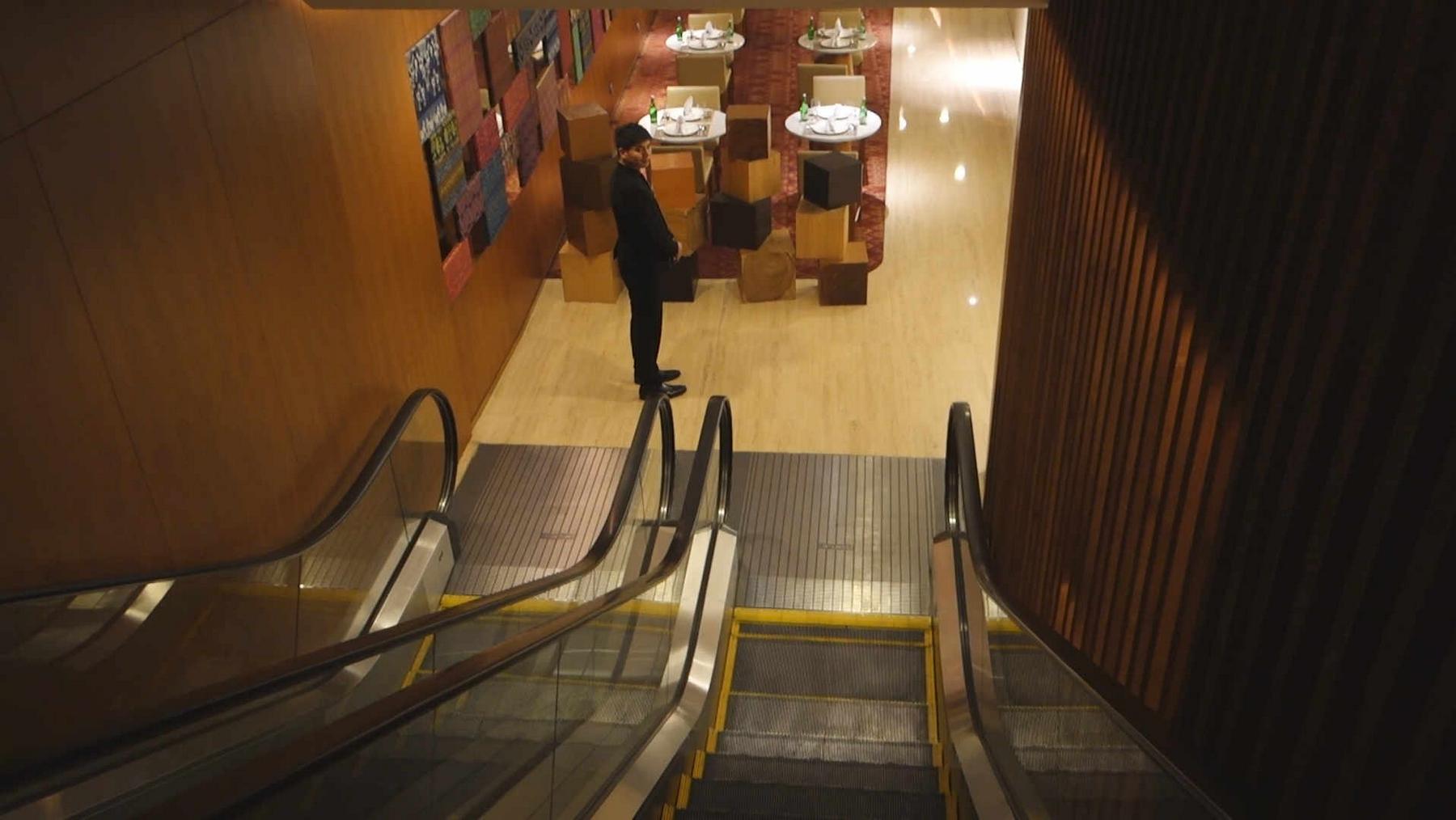 A person stands in a lounge area with dining tables and stacked boxes, viewed from the top of an escalator.