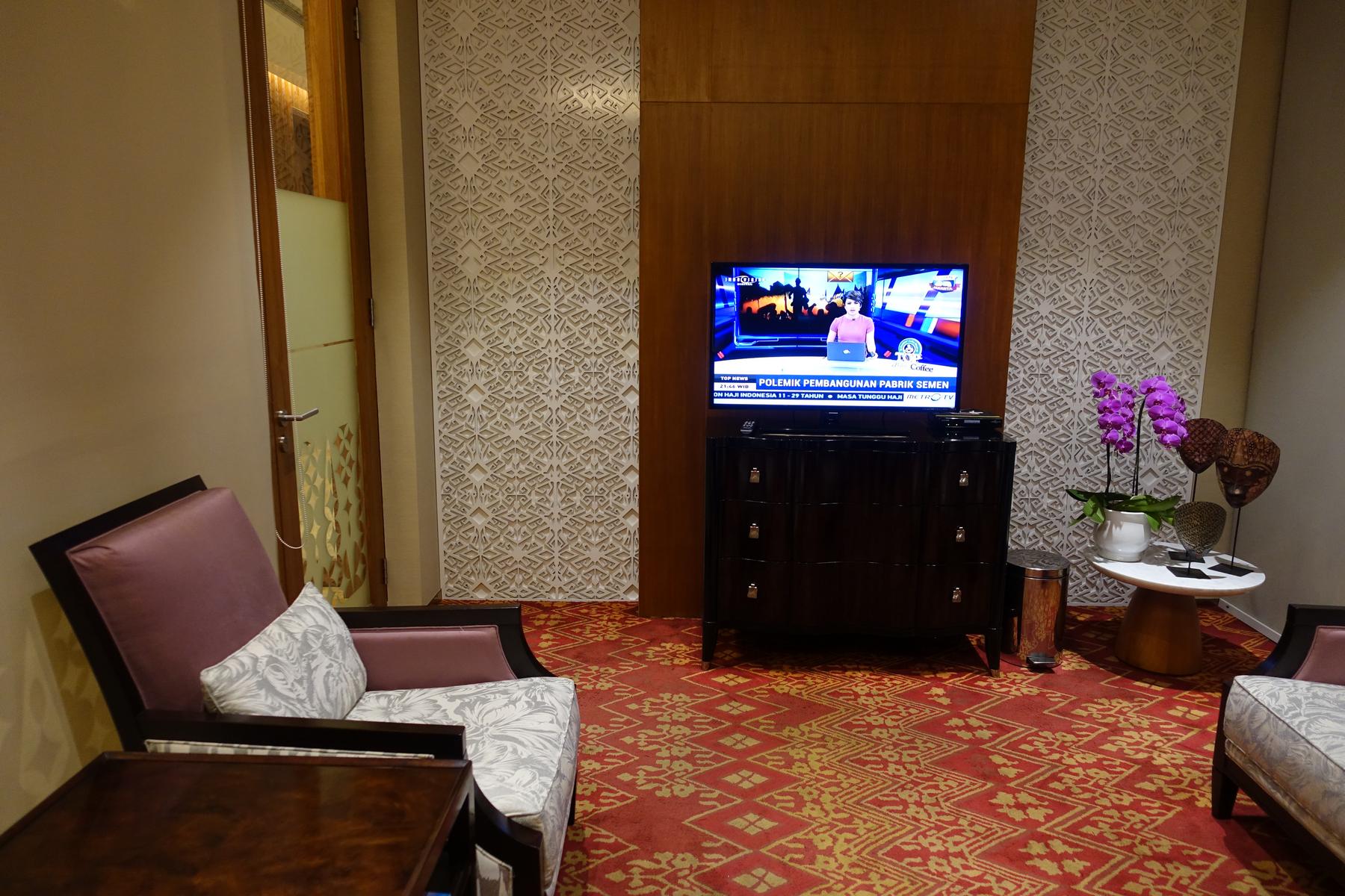 An elegant lounge area with a TV, plush armchairs, ornate wall paneling, and a red patterned carpet.