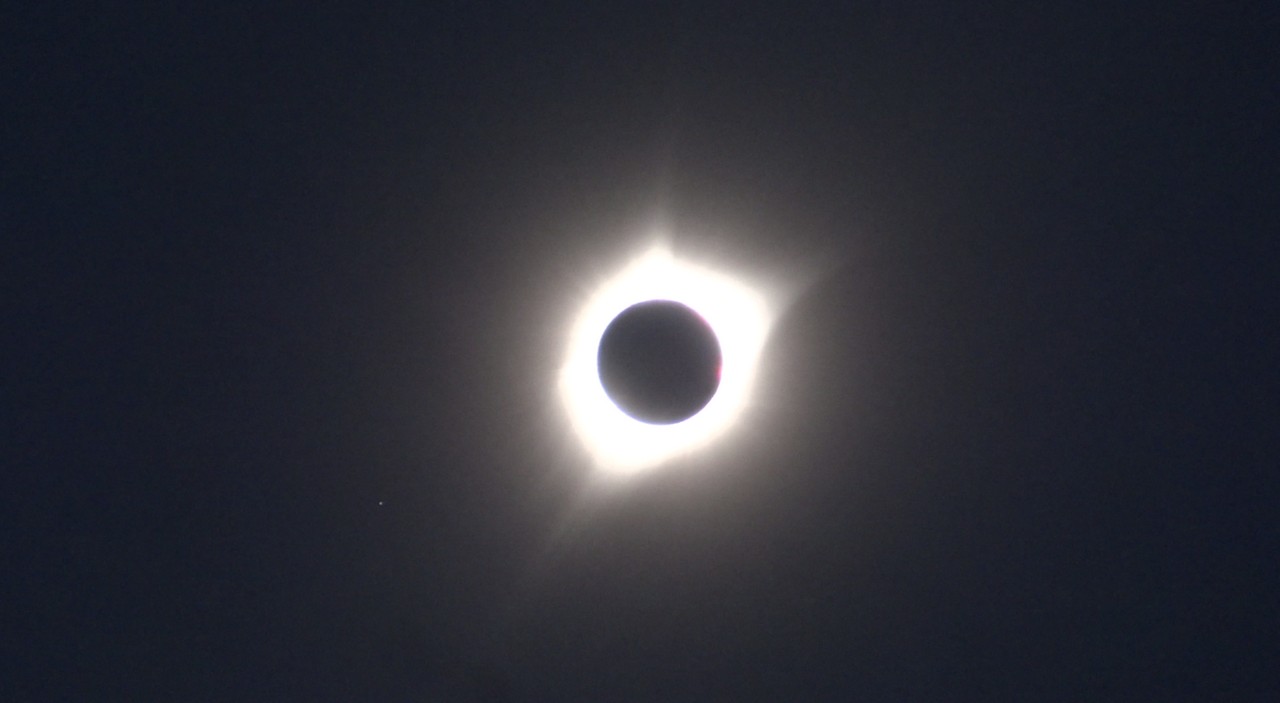 A total solar eclipse showing the sun's brilliant white corona around the dark moon, with a bright star or planet visible.
