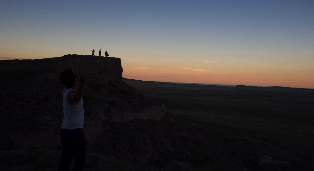 Family members in silhouette watch the twilight sky from a Nebraska bluff, one with hands raised.