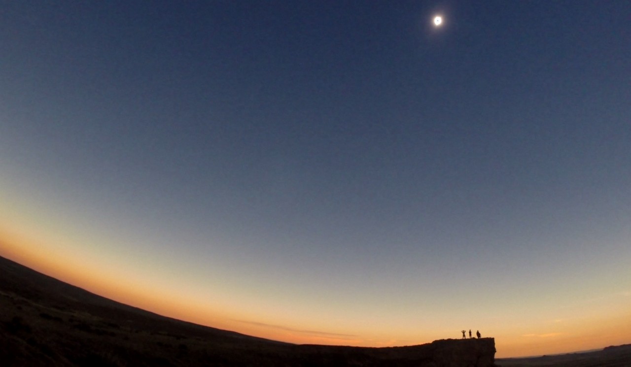 A total solar eclipse is visible in the twilight sky above a family silhouetted on a cliff.
